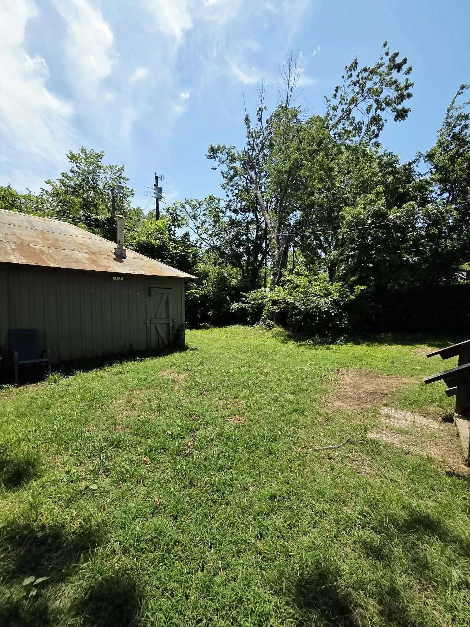 Spacious grassy area featuring a utility building with a metal roof and board siding, surrounded by mature trees
