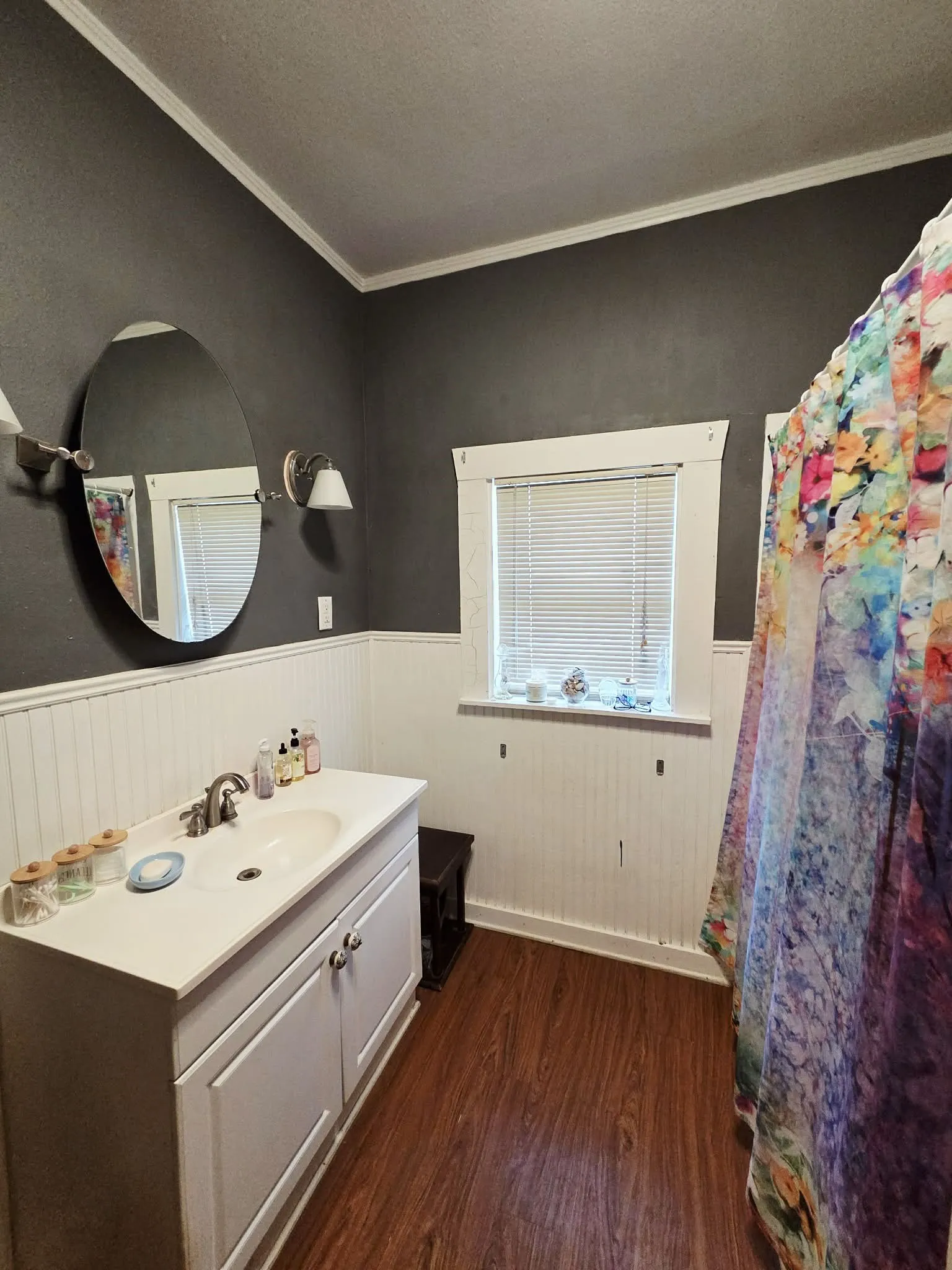 Bathroom featuring wood-finish flooring, a white vanity with an integrated sink, a window with blinds, and white wainscoting