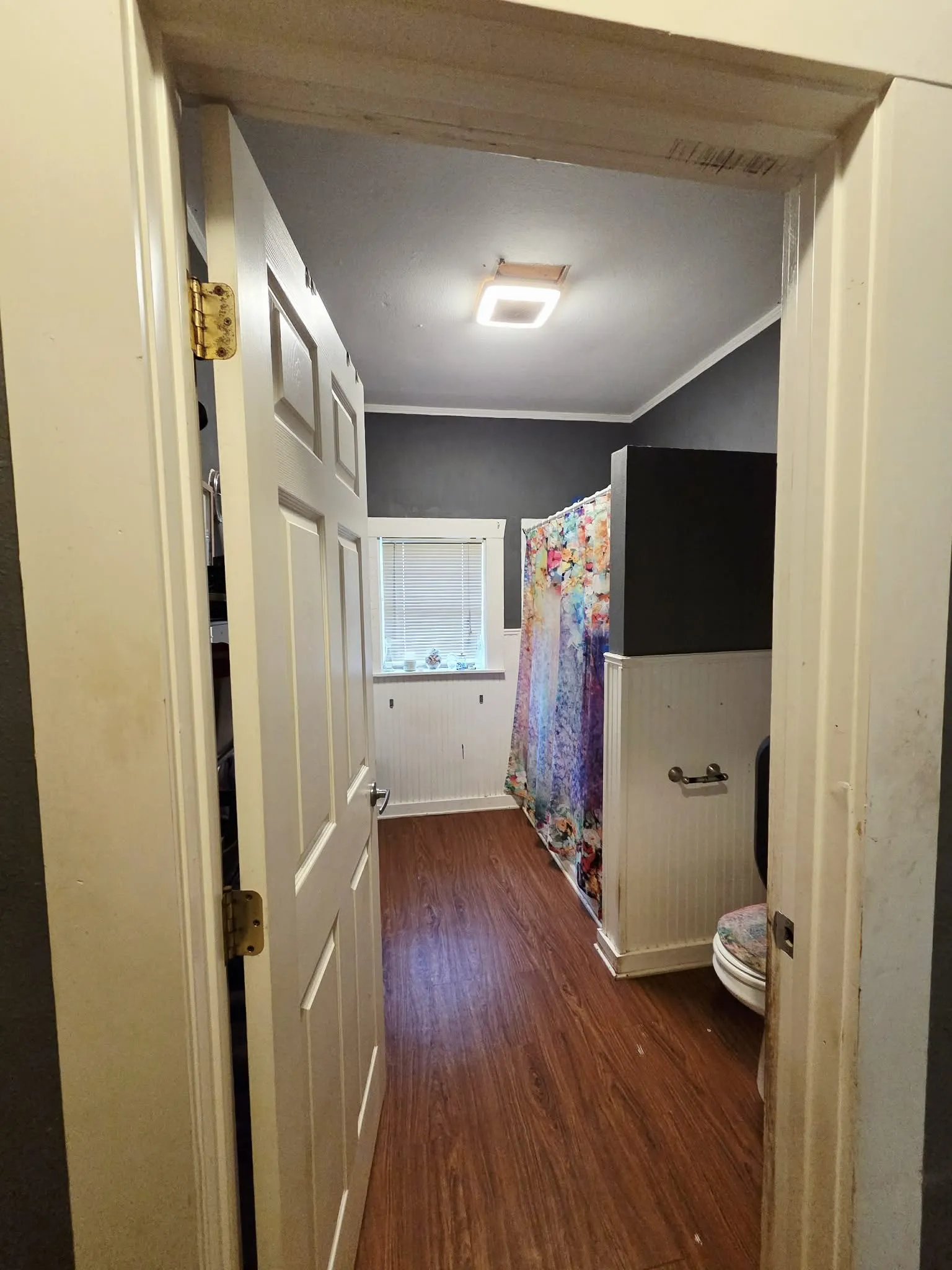 Bathroom featuring wood-finish flooring, dark wall paint, white wainscoting, and a window with blinds