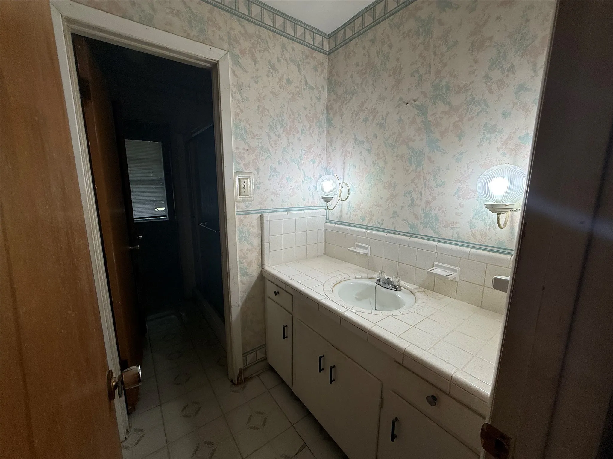 Bathroom vanity featuring a tiled countertop with an integrated sink, dual wall-mounted sconces, and a patterned wallpaper accent