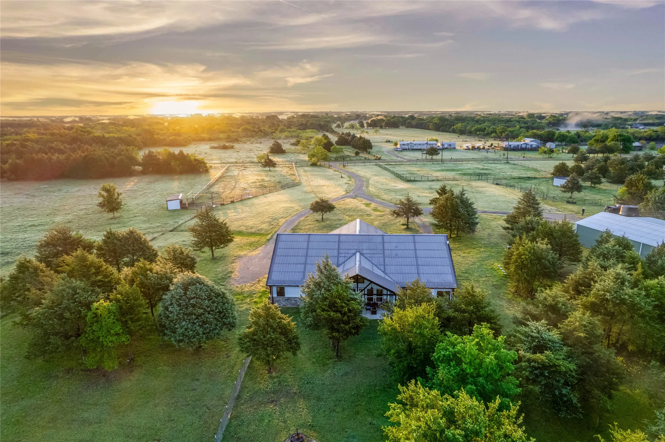 Expansive rural property featuring a prominent main residence with a dark-paneled roof, surrounding green foliage, and multiple fenced pasture areas