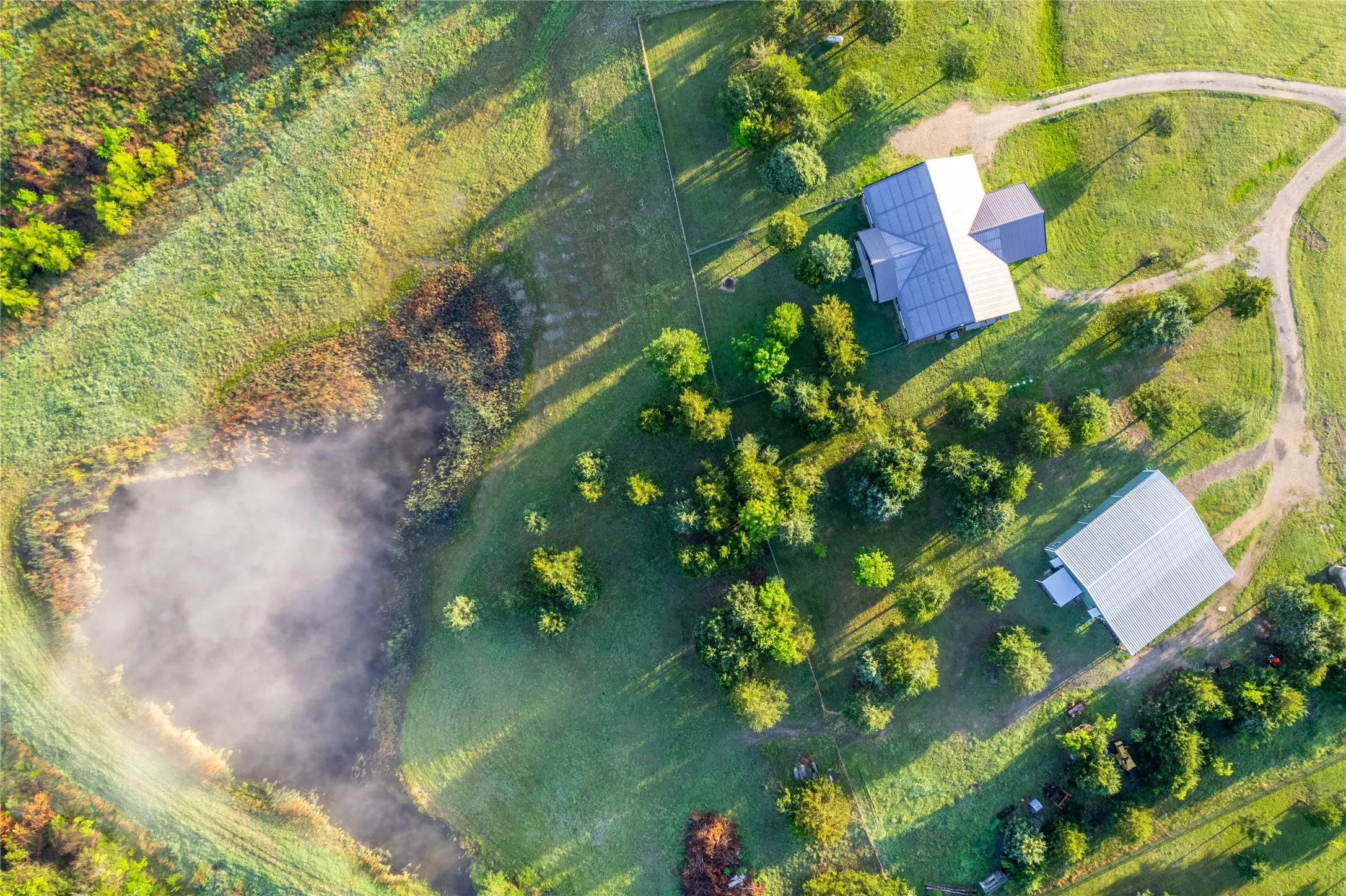 Expansive property featuring a private pond, main residence with a metal roof, secondary building with a metal roof, winding gravel driveways, and mature landscaping