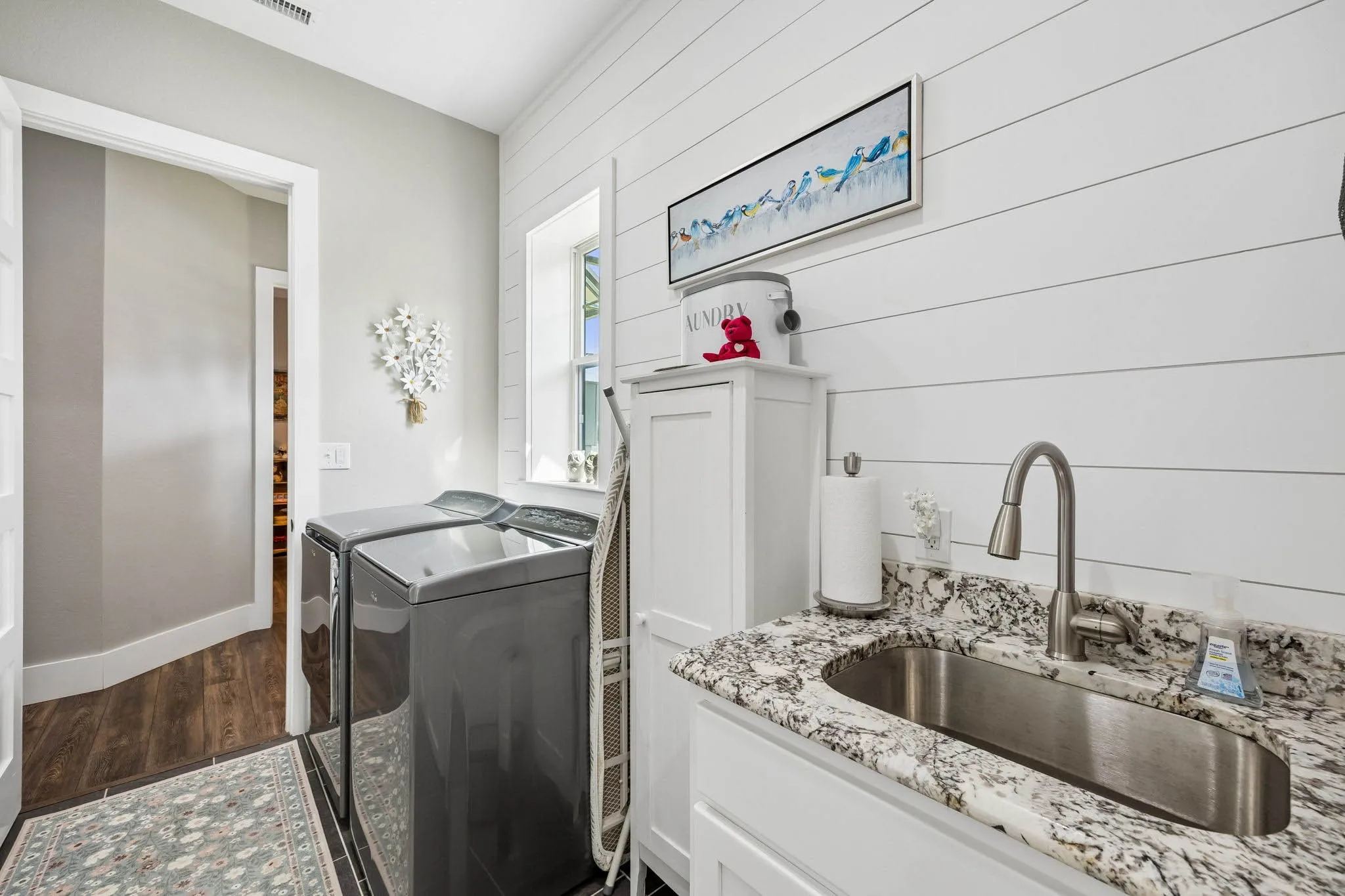 Laundry room featuring white shiplap walls, a window, a white base cabinet with a granite-style countertop, and an undermount stainless steel sink with a gooseneck faucet