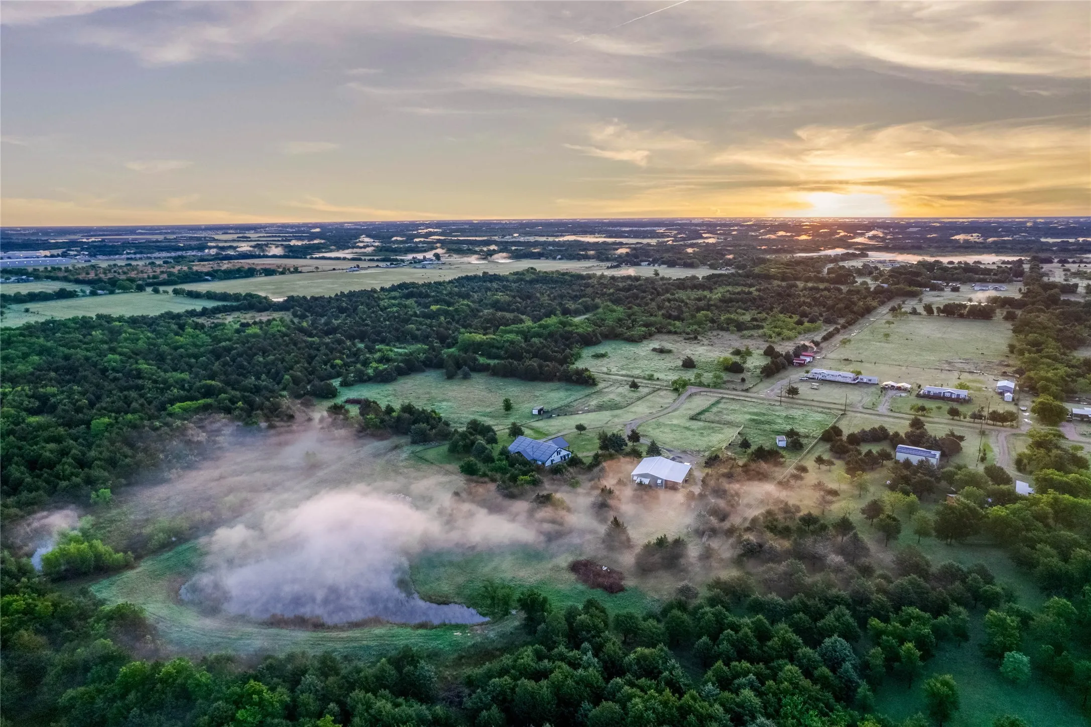 Expansive rural landscape featuring a private pond, abundant mature trees, open clearings, and multiple residential structures
