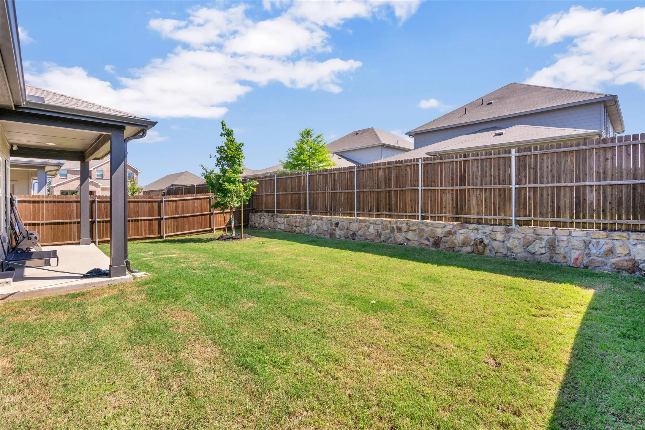 Expansive grass yard featuring a stone retaining wall, mature privacy fencing, and a covered concrete patio