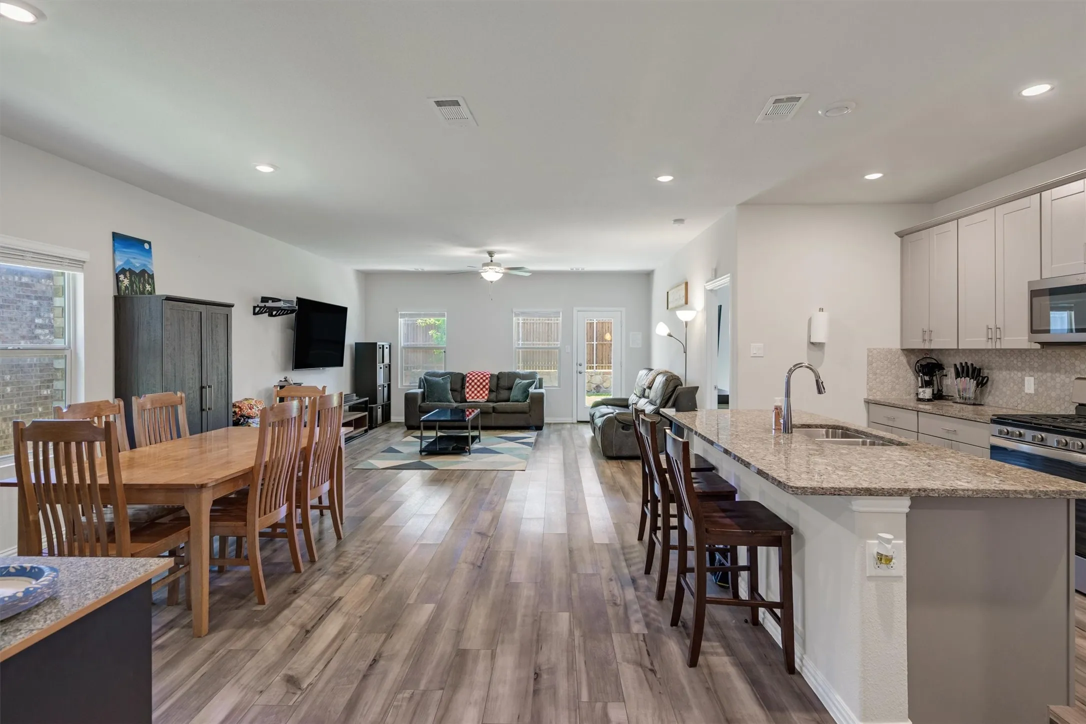 Open-concept living space featuring wood-finish flooring, a kitchen island with stone countertop and integrated sink, light-toned cabinetry, stainless steel appliances, and recessed lighting