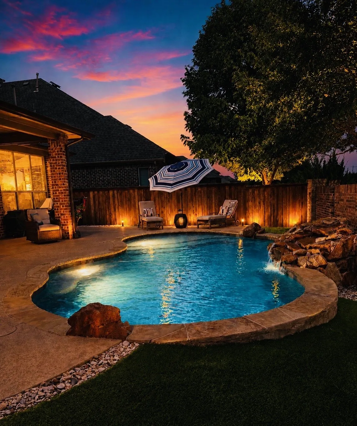 Freeform swimming pool with integrated rock waterfall feature, ambient landscape lighting, concrete decking, and a privacy fence