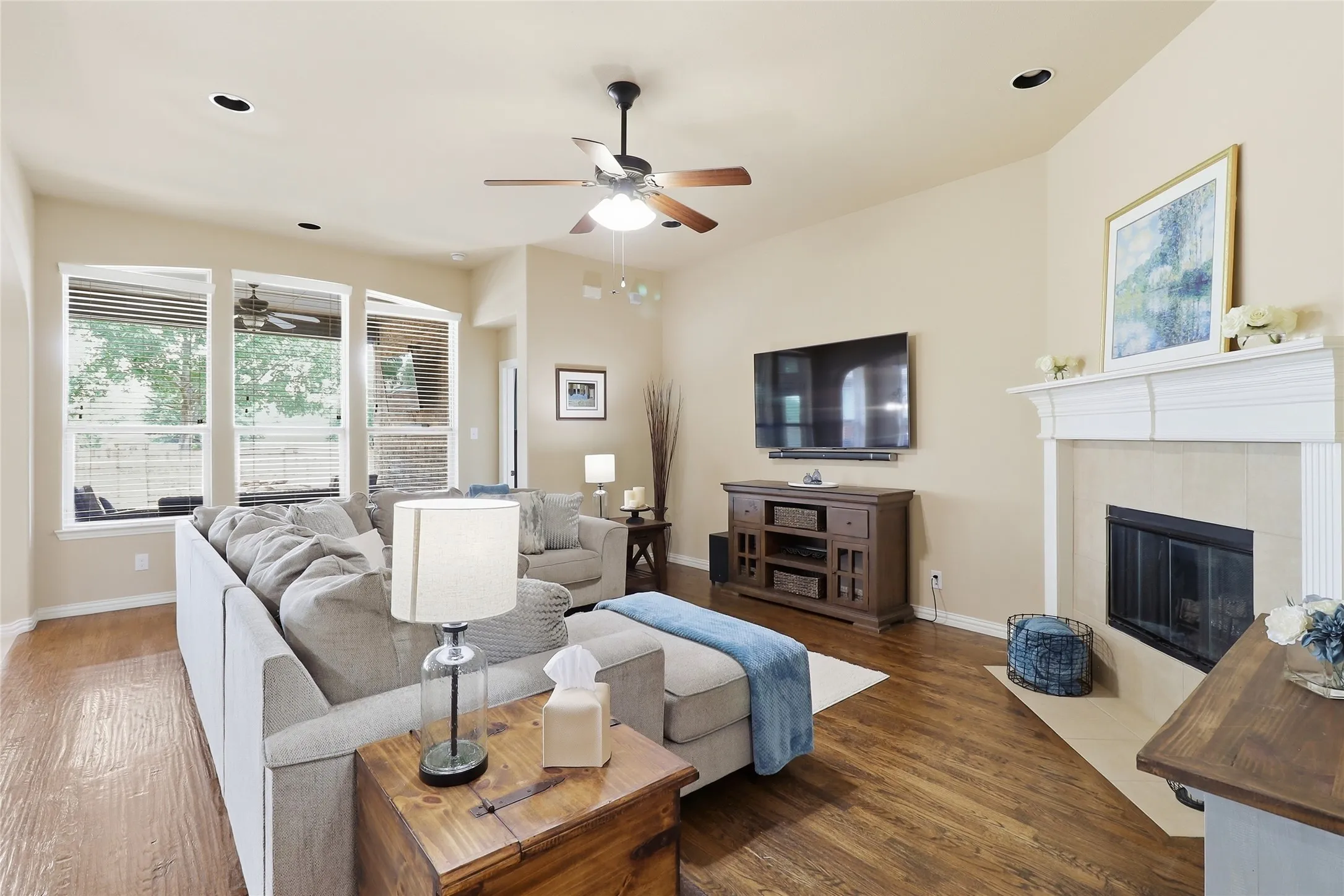 Living room featuring wood-finish flooring, a fireplace with a light-colored tile surround and white mantel, a ceiling fan, and recessed lighting