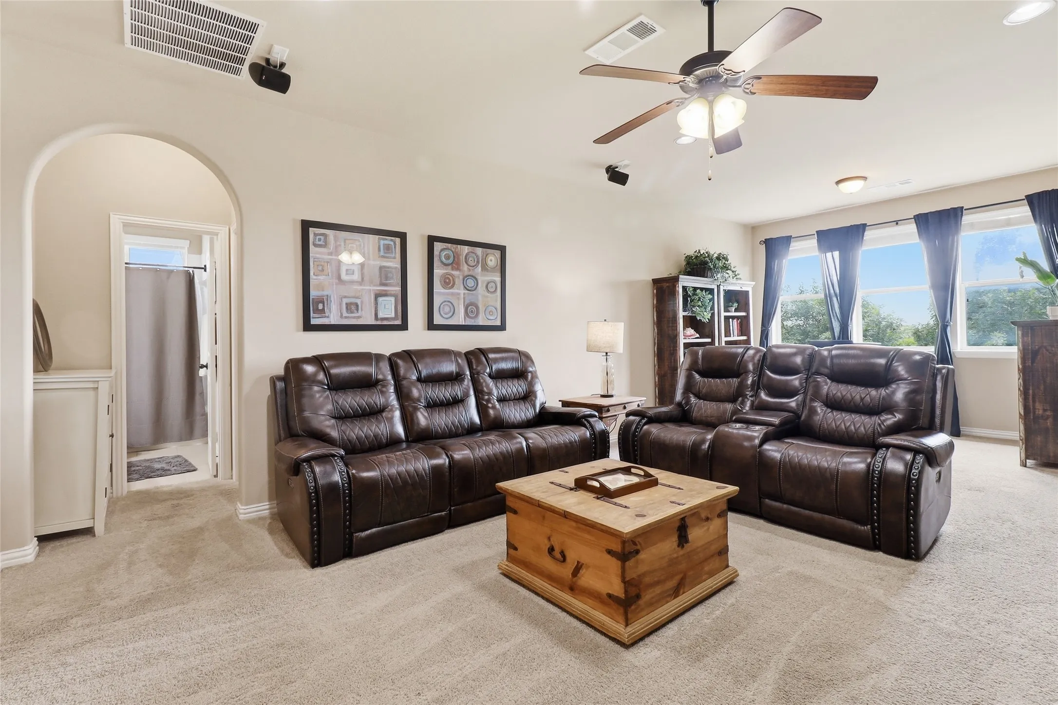Spacious carpeted room featuring a ceiling fan with integrated lighting, recessed overhead speakers, multiple windows, and an arched doorway