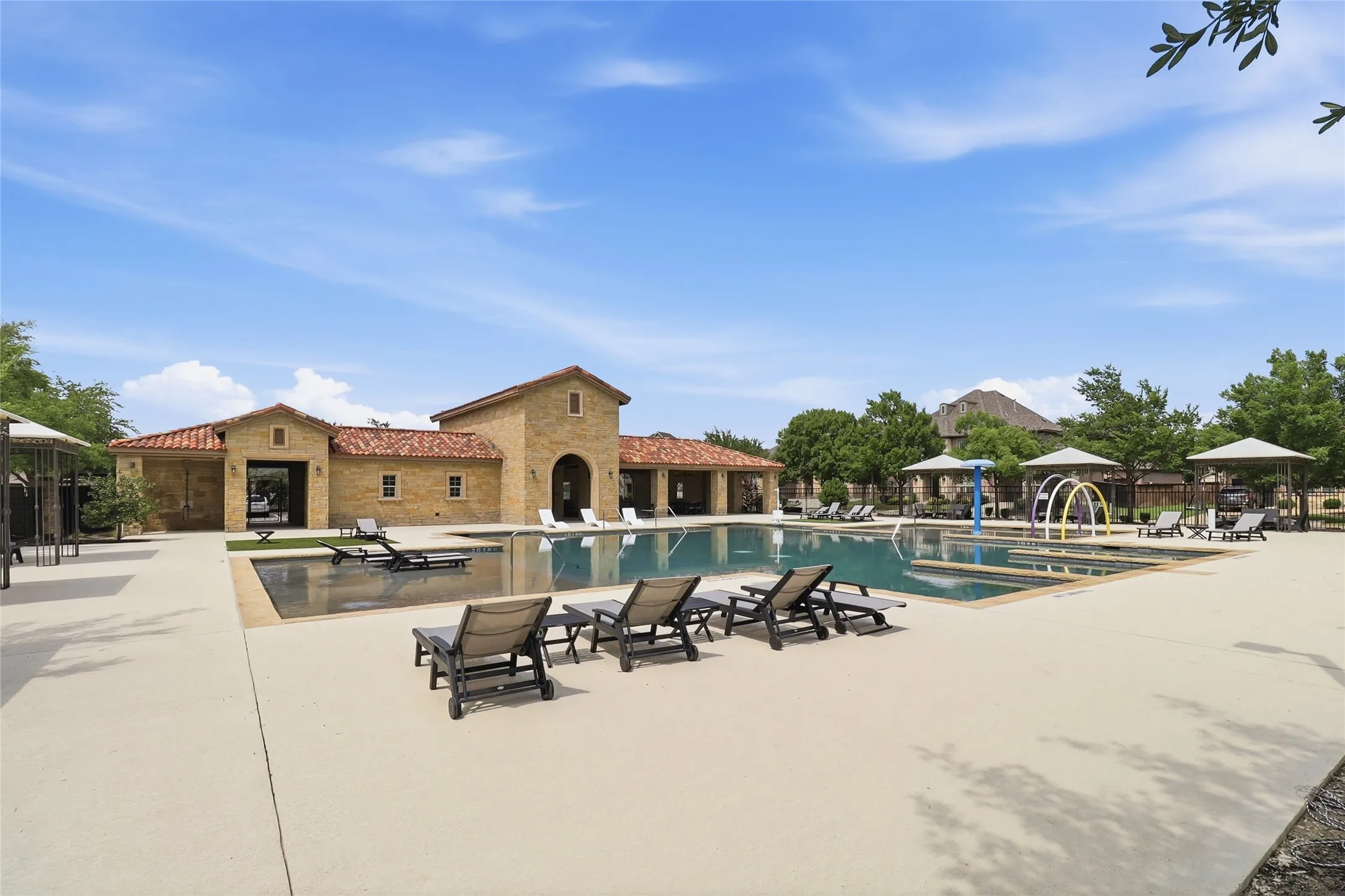 Resort-style pool area featuring a stone-clad building with a tile roof, multiple chaise lounges, a splash pad with water features, and shaded seating areas