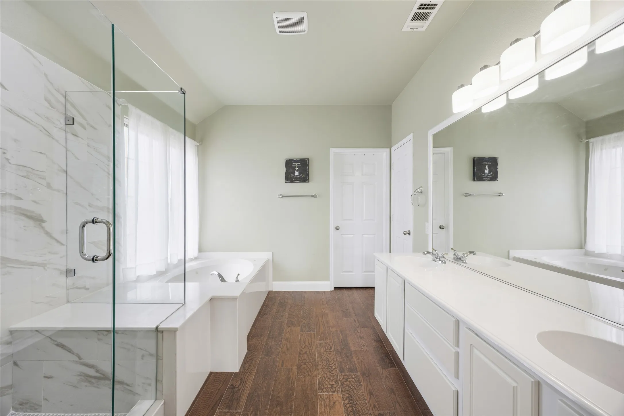Spacious bathroom featuring a glass-enclosed shower with marble-look tile, a built-in soaking tub, a long double vanity with integrated sinks, and wood-finish flooring