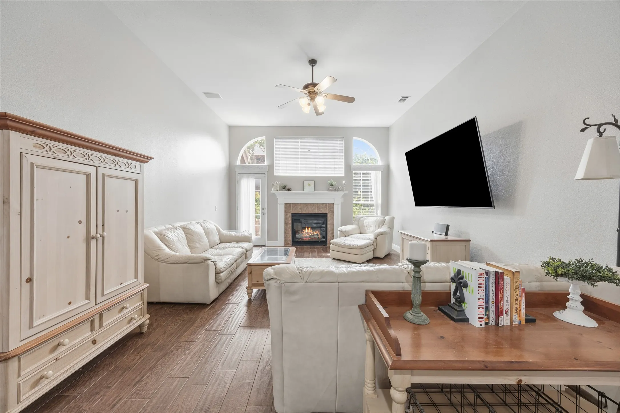 Living room featuring wood-finish flooring, a fireplace with a stone surround and white mantel, and arched windows