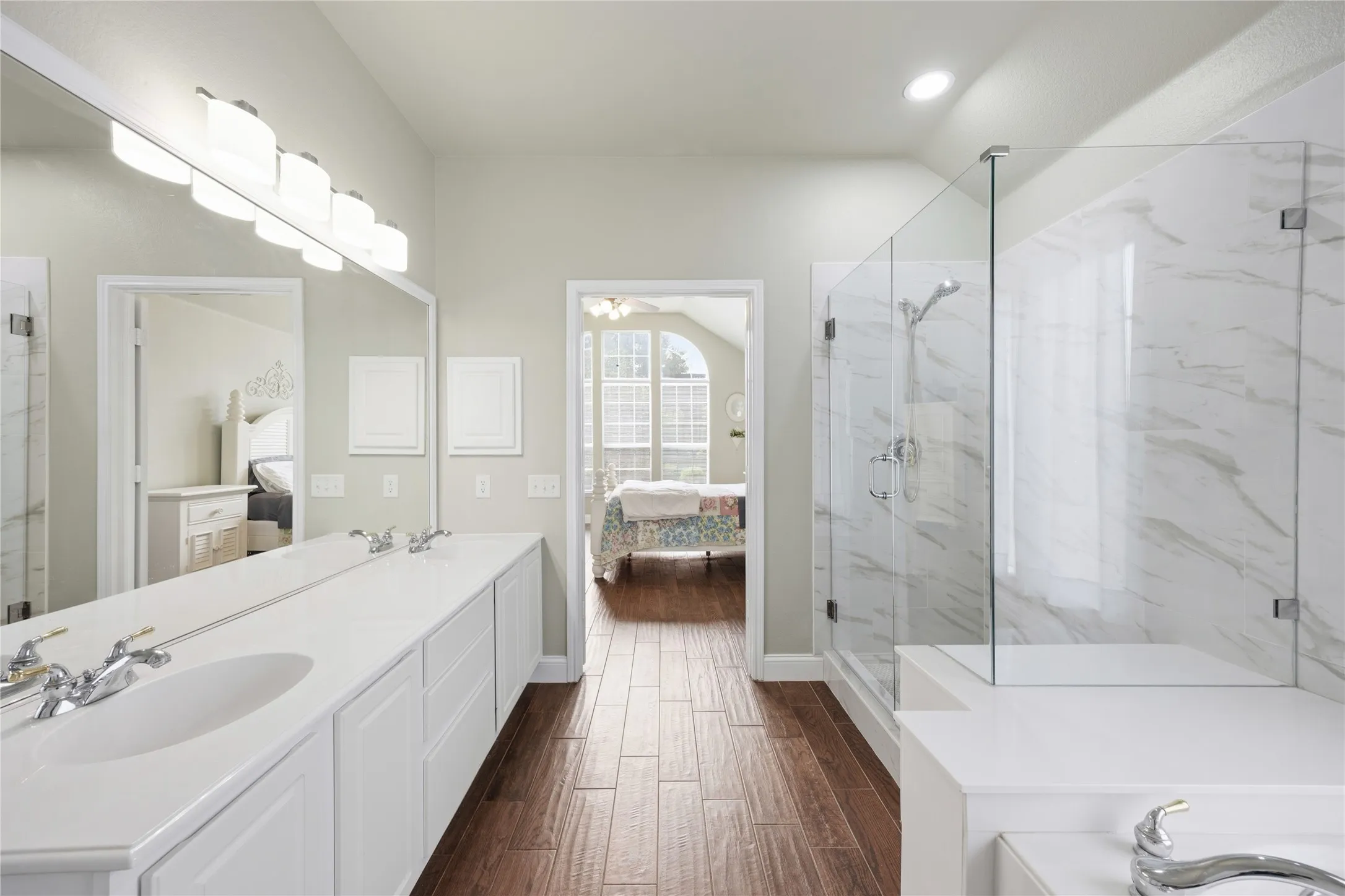 Wood-finish flooring throughout, featuring a dual-sink vanity with white cabinetry, a full-width mirror, and a glass-enclosed shower with white marble-patterned tile surround