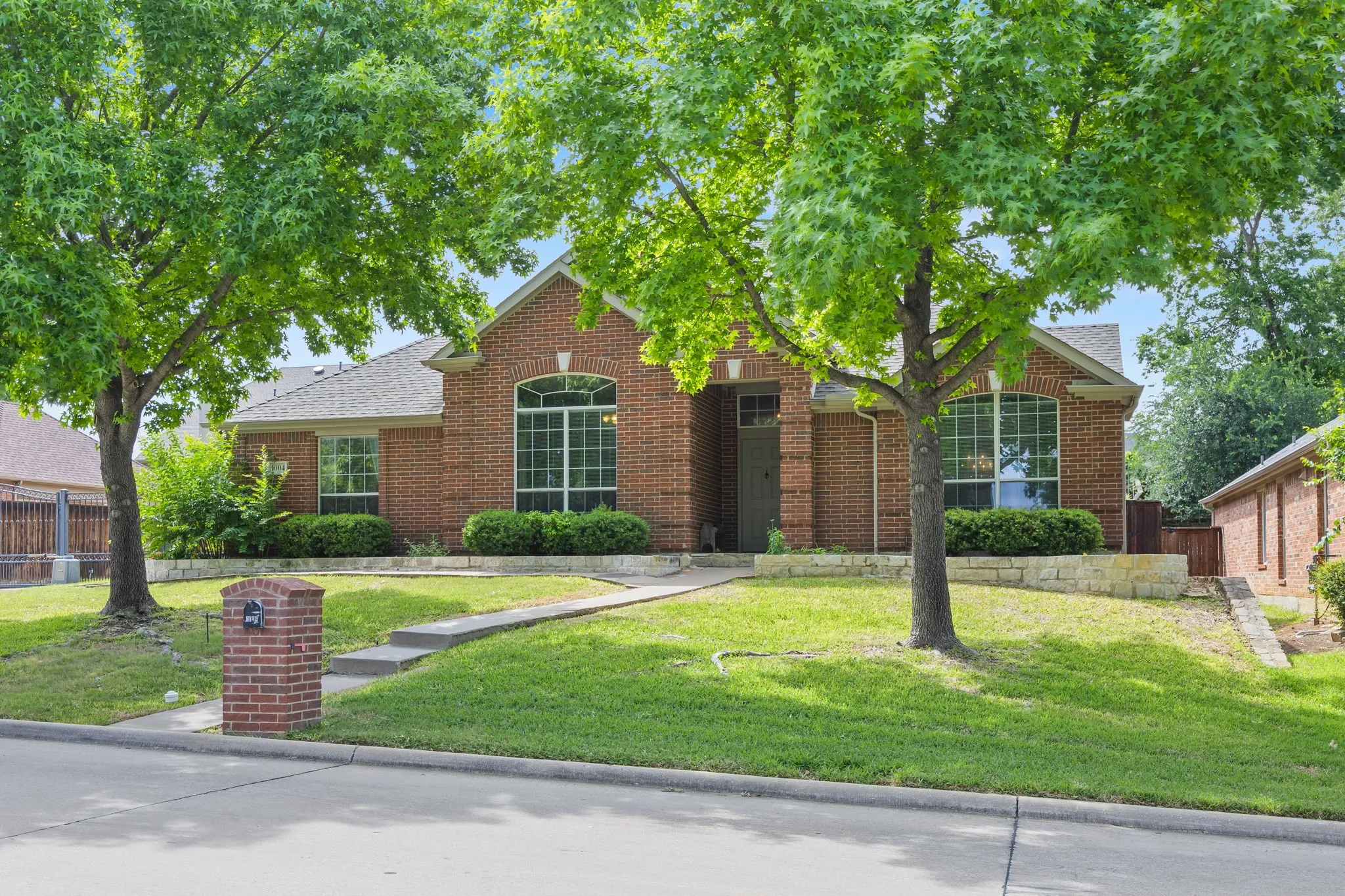 Traditional brick exterior featuring an arched window, a gabled roofline, mature trees, a manicured lawn, and a stone retaining wall