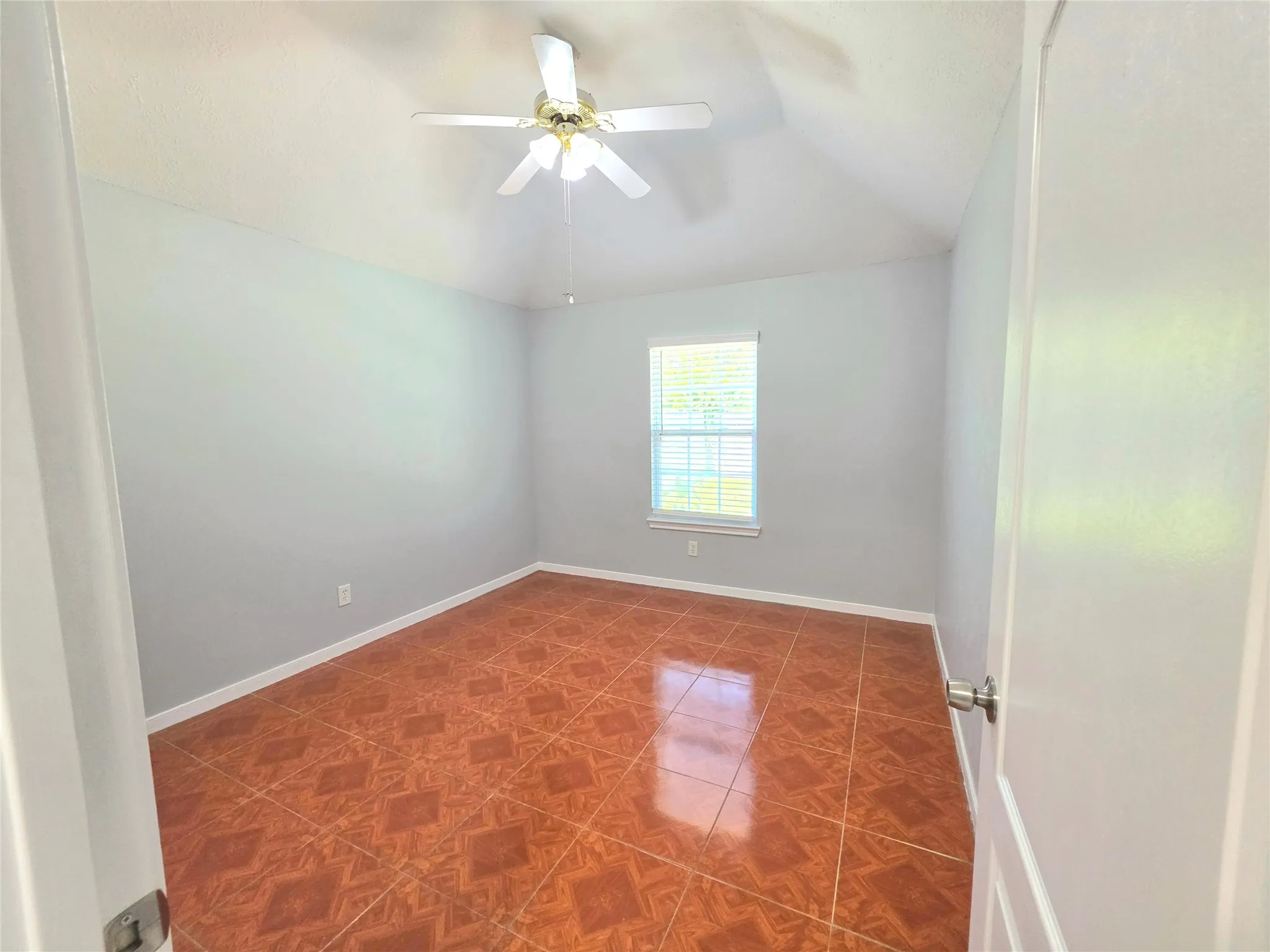 Spacious room featuring patterned terracotta flooring, a white ceiling fan with integrated lighting, a single window with blinds, and light gray wall paint