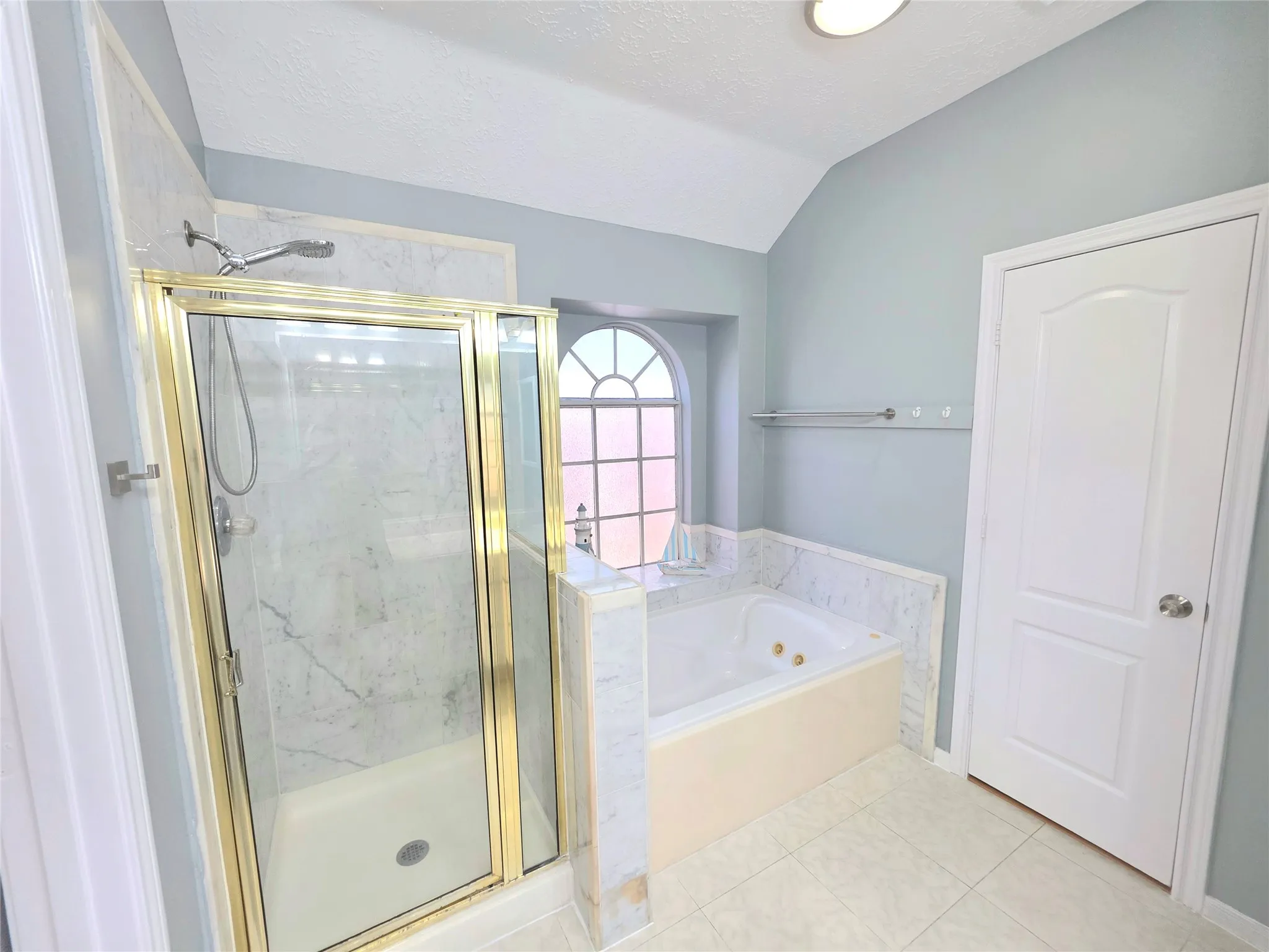 Bathroom featuring a built-in jetted tub, a separate shower with a gold-finish frame, and an arched window