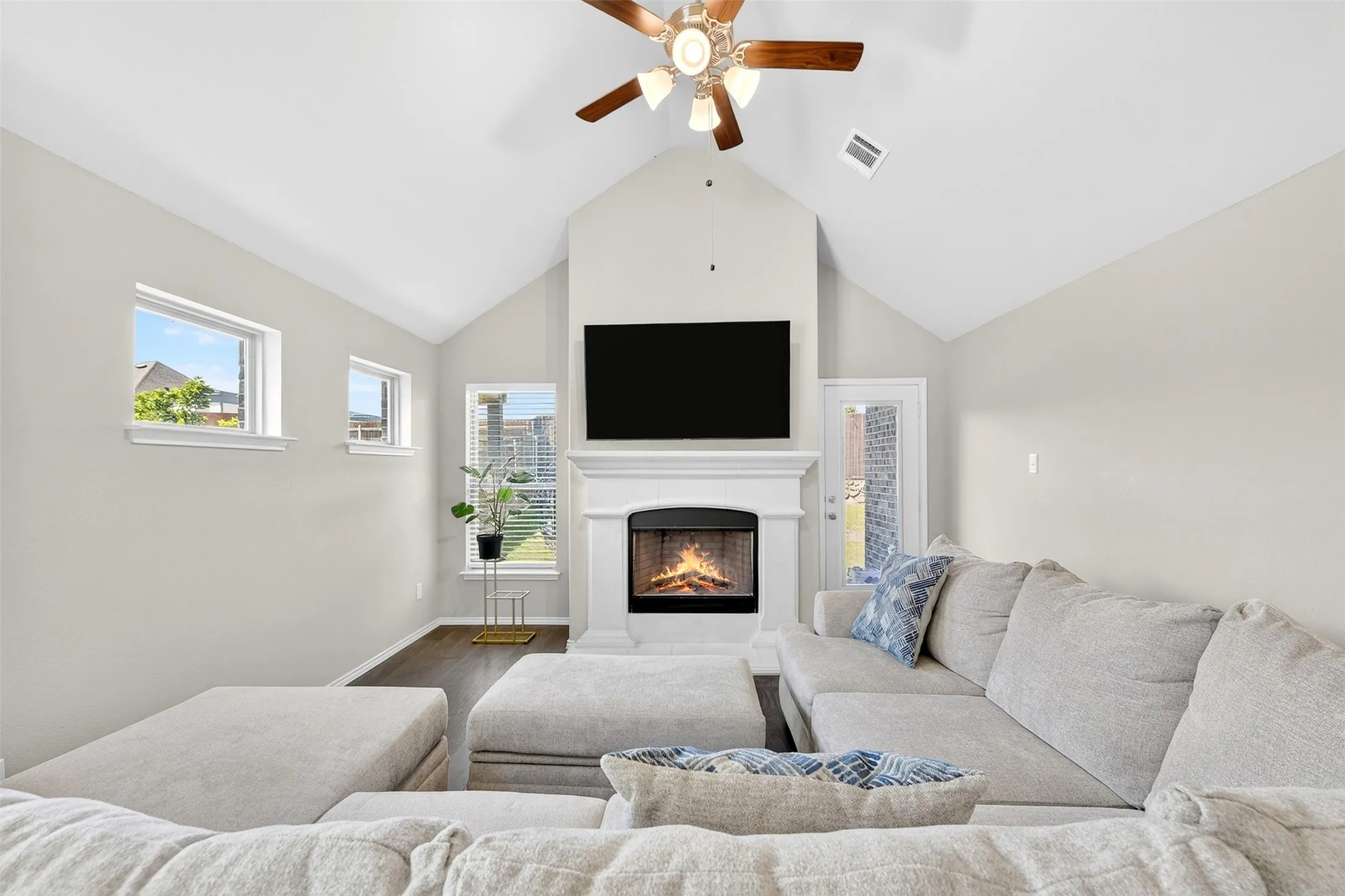 Vaulted ceiling living area featuring a fireplace with a white mantel, dark wood-finish flooring, multiple windows, and a ceiling fan