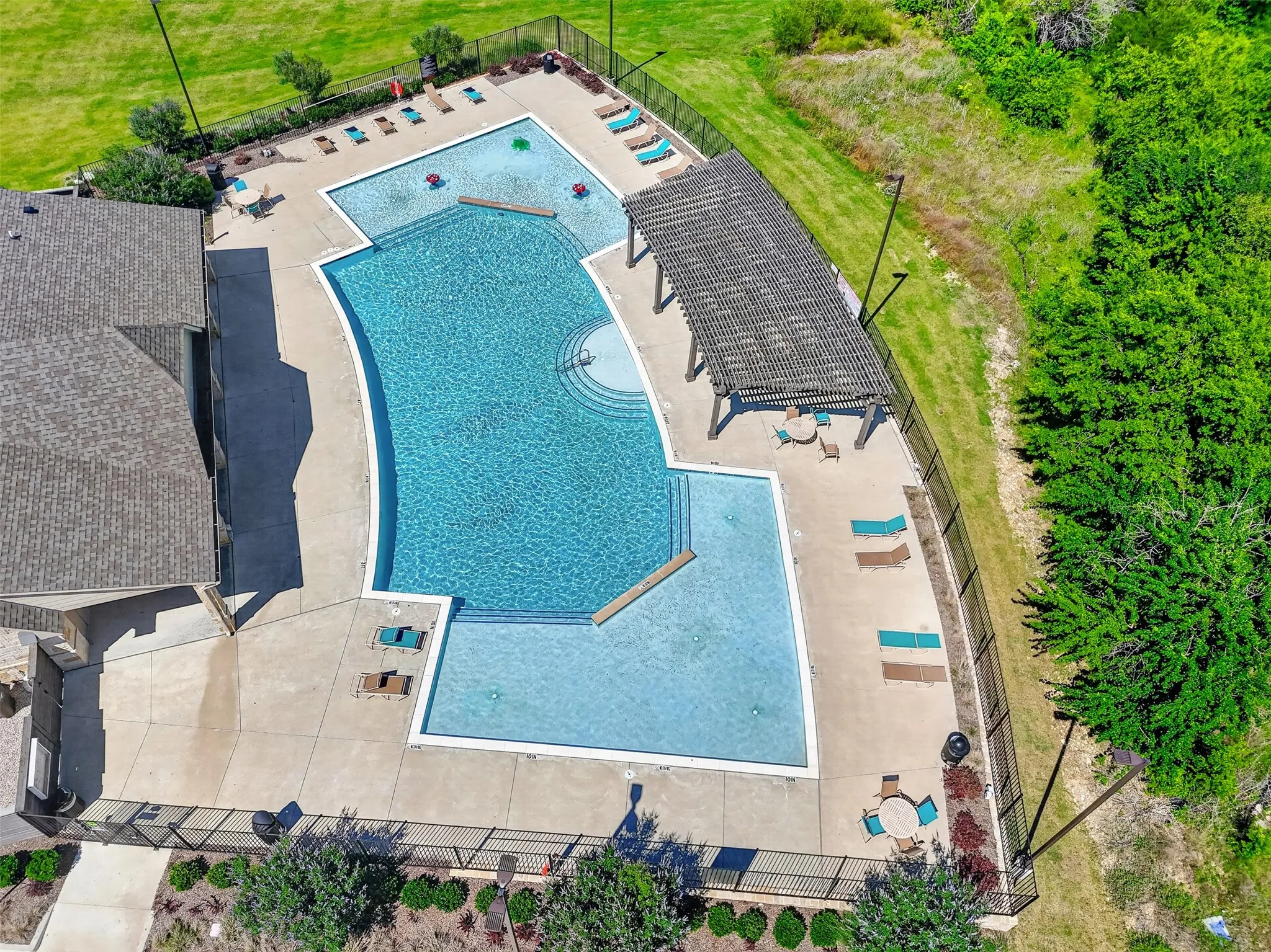 Resort-style pool area featuring multiple swimming sections, a built-in pergola, extensive concrete decking, surrounding black metal fencing, and integrated landscaping