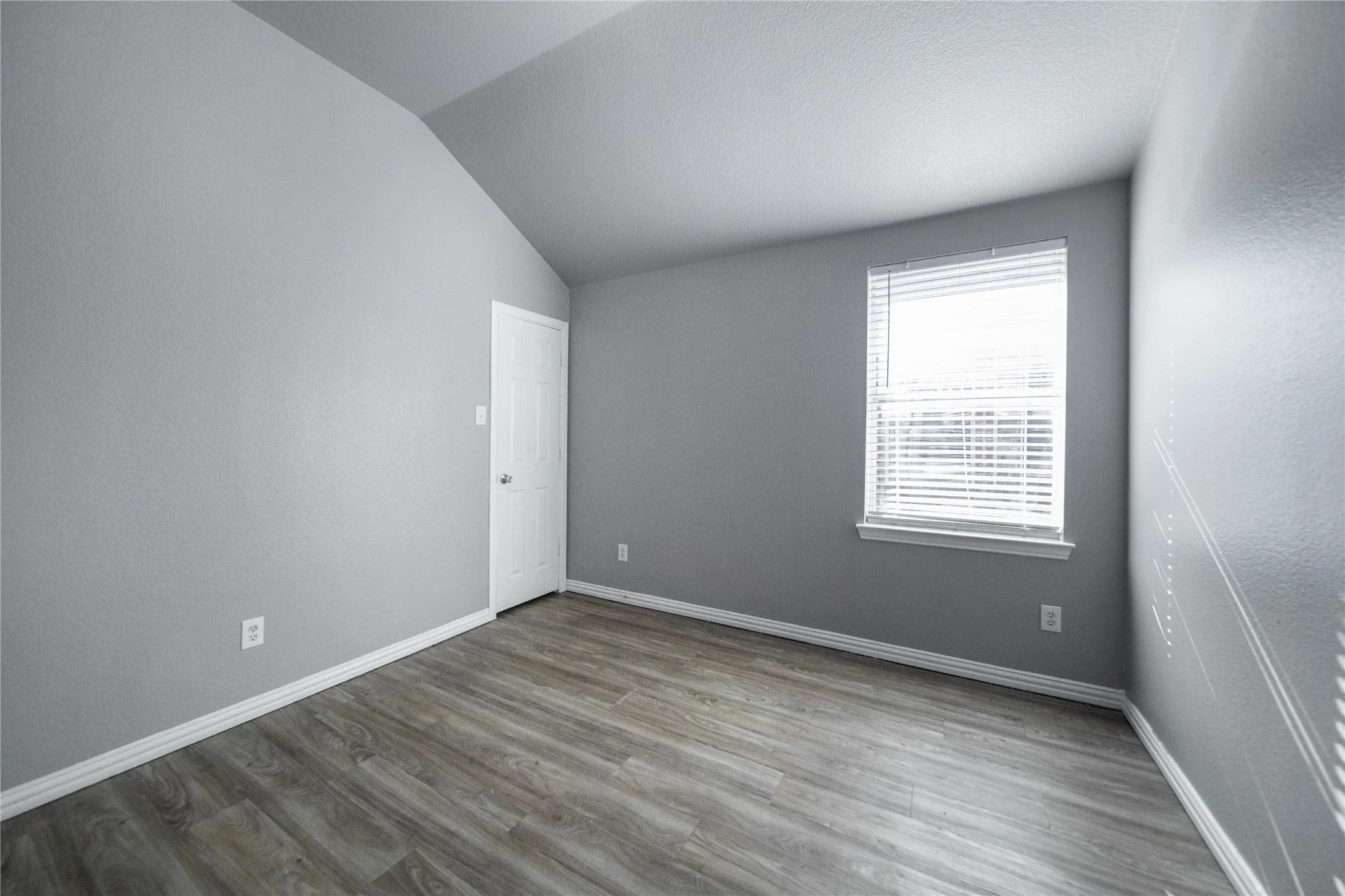 Room featuring wood-finish flooring, white baseboards, a white paneled door, and a window with blinds