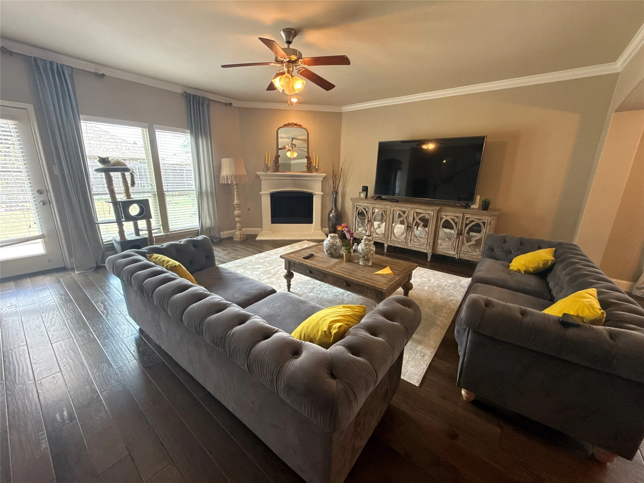 Spacious living area featuring rich wood-finish flooring, a corner fireplace with a white mantel, and a ceiling fan with integrated lighting