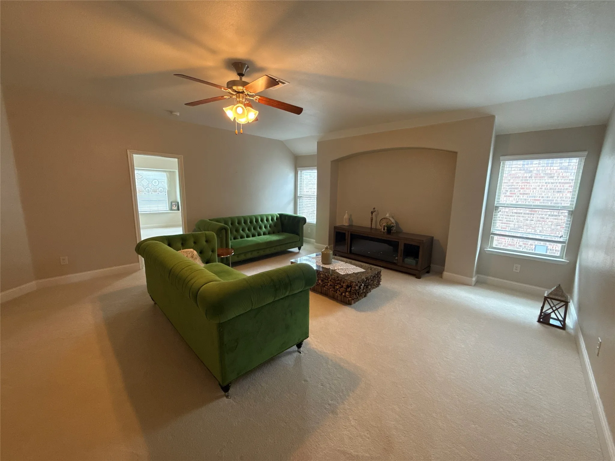 Spacious living area featuring neutral carpeting, a built-in arched alcove, recessed windows with blinds, and a ceiling fan with integrated lighting