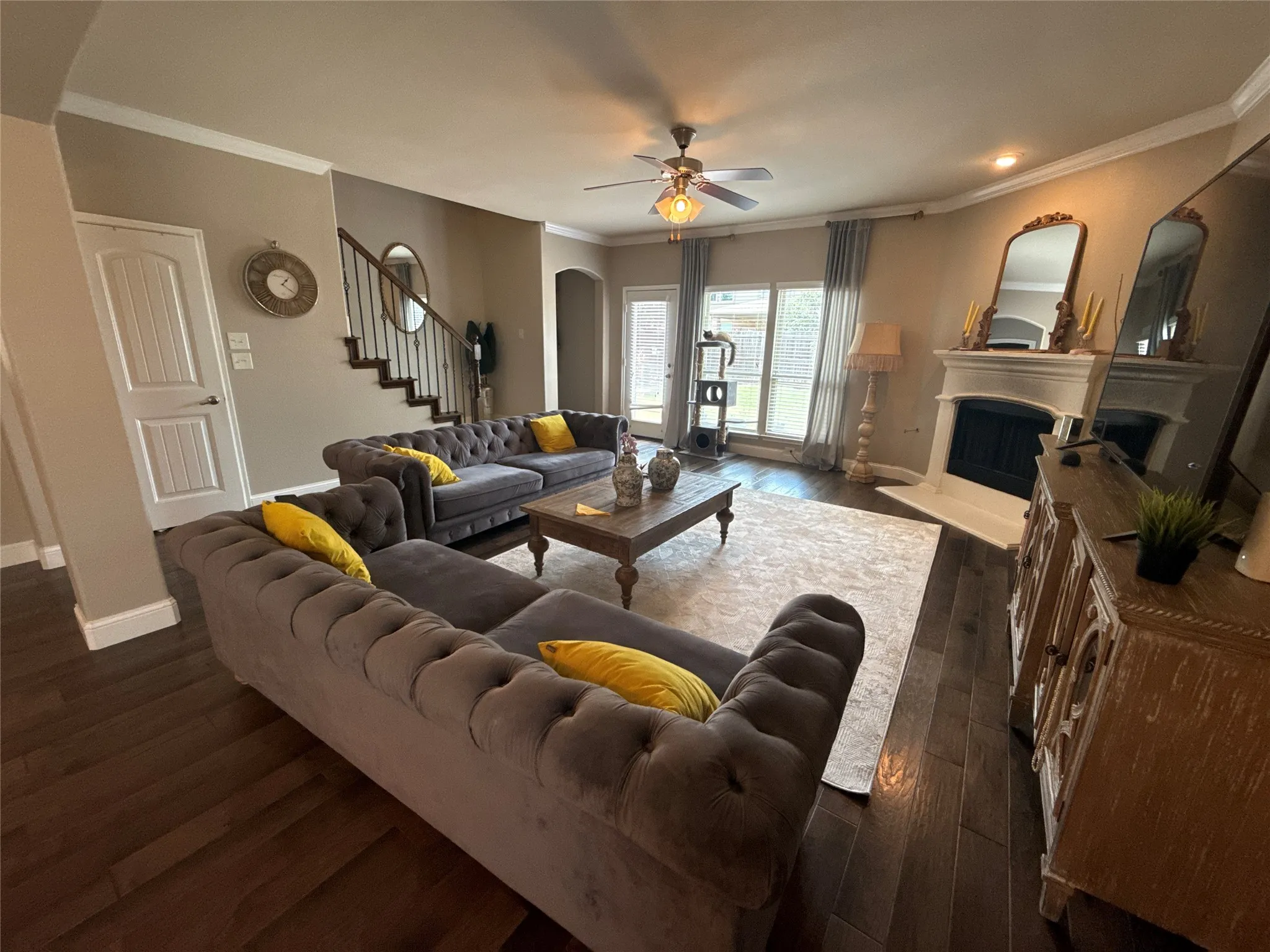 Spacious living area featuring dark wood-finish flooring, a white fireplace surround, and a ceiling fan with integrated lighting