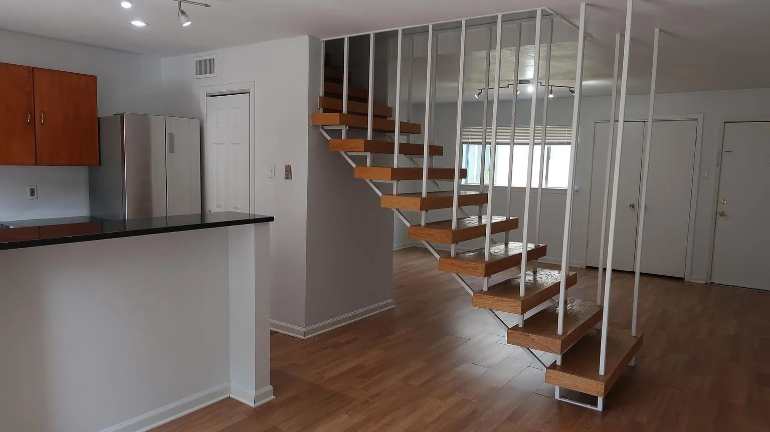 Open-concept living area featuring wood-finish flooring, a contemporary floating staircase with white railings, and a kitchen peninsula with a dark countertop