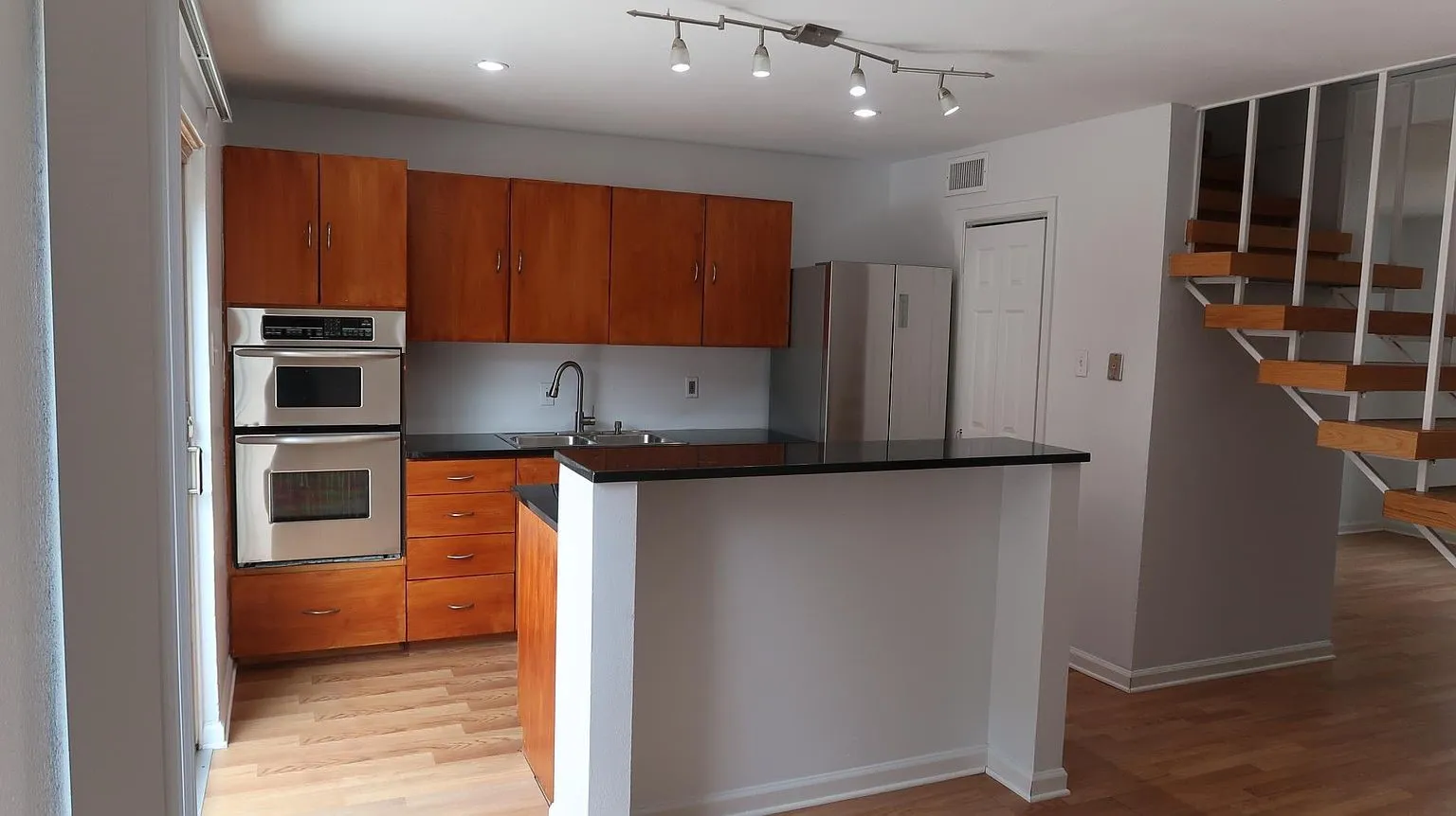 Kitchen featuring wood-finish cabinetry, built-in stainless steel microwave, oven, black countertops, and a track lighting fixture