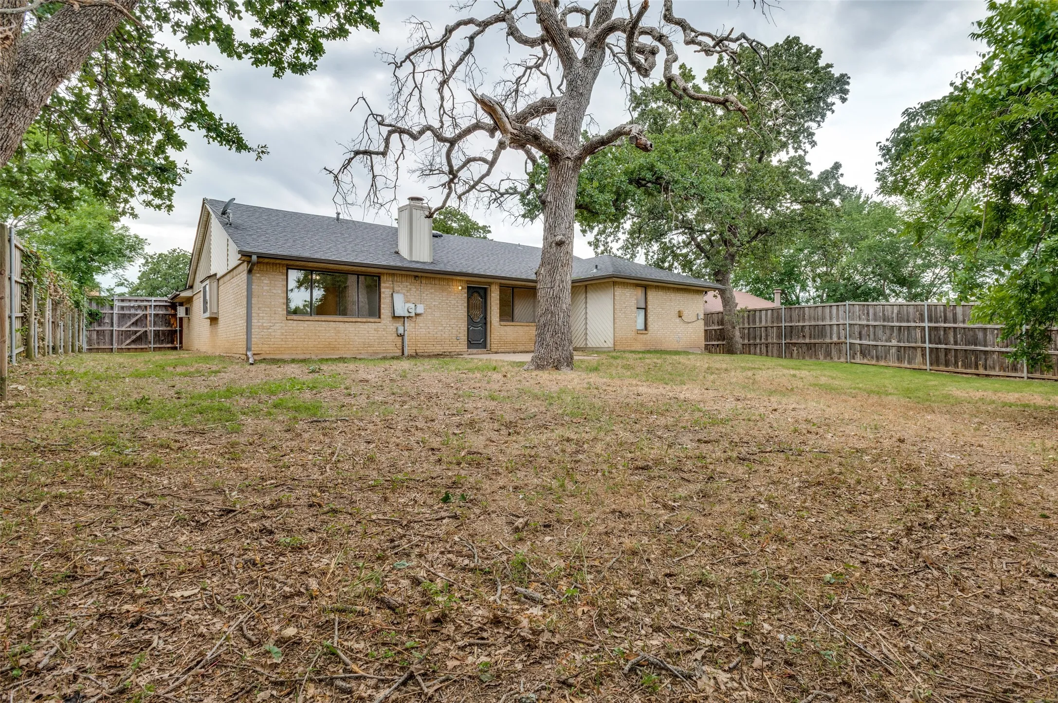 Expansive backyard featuring mature trees, a full privacy fence, and a brick exterior home with a dark shingle roof