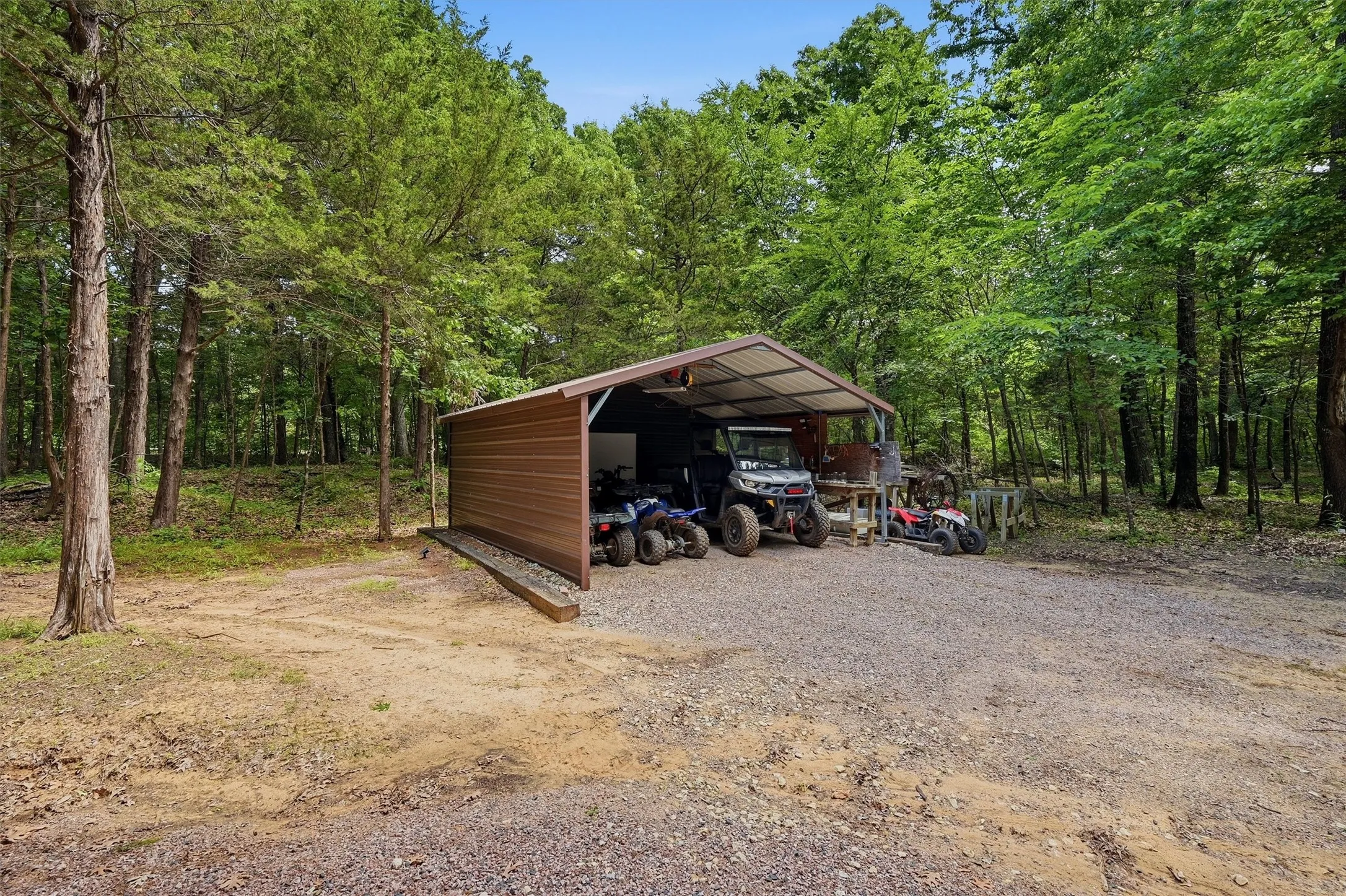 Metal carport structure with a gravel base, surrounded by mature trees