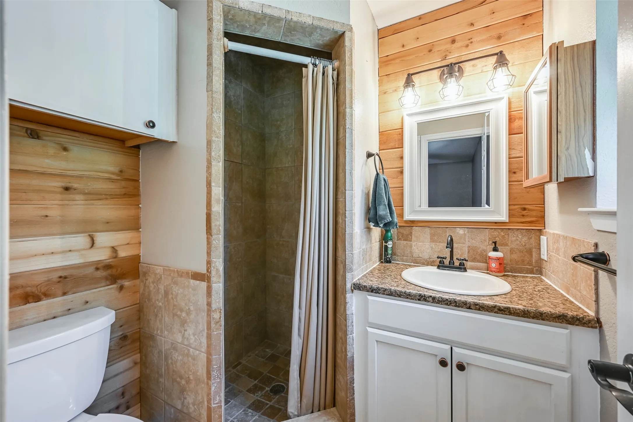 Bathroom featuring a tiled shower, wood-finish accent wall, single vanity with a built-in sink, countertop, and mounted mirror with overhead lighting