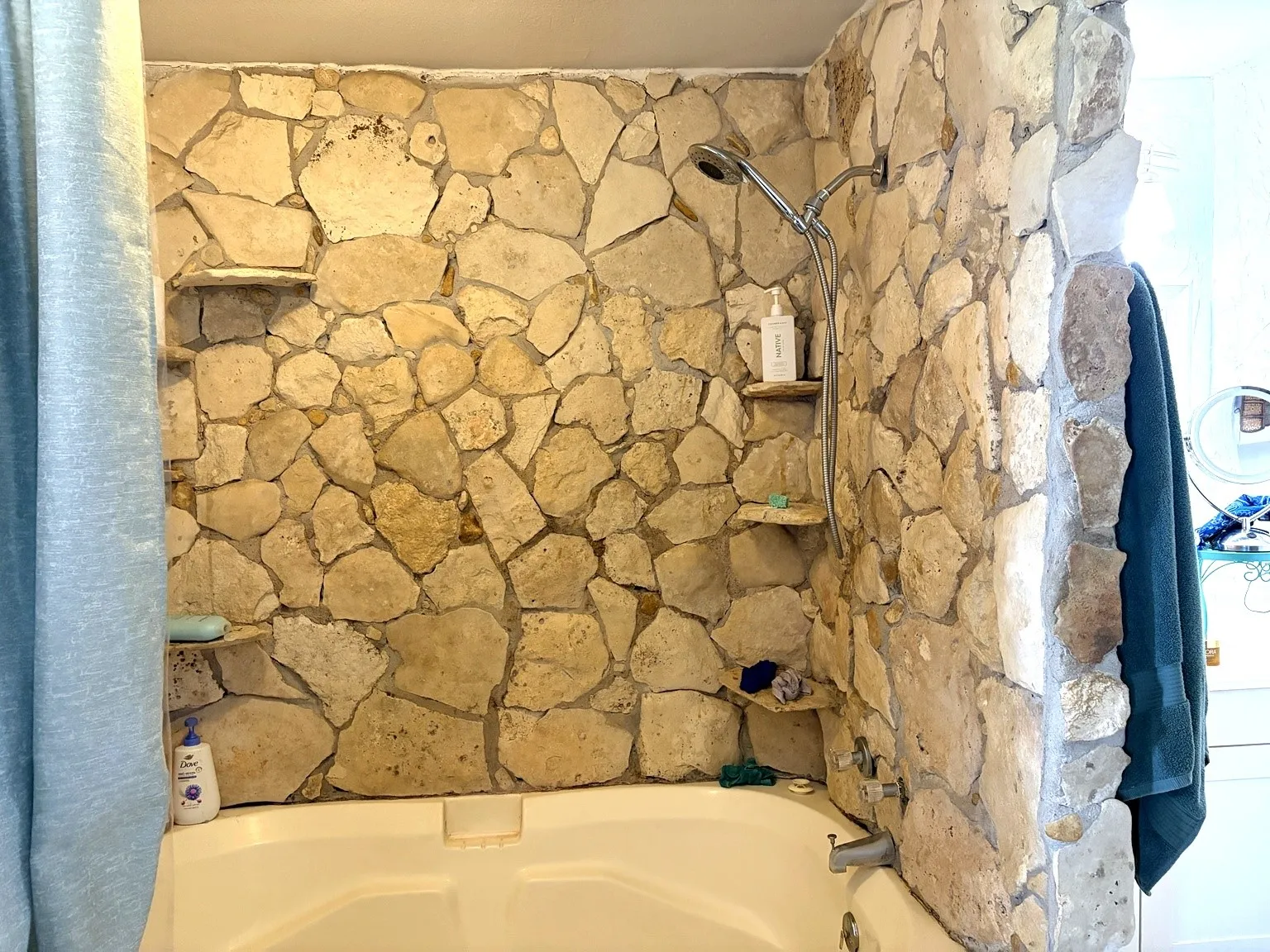 Bathtub with stone-clad walls, featuring a handheld showerhead and built-in stone shelving