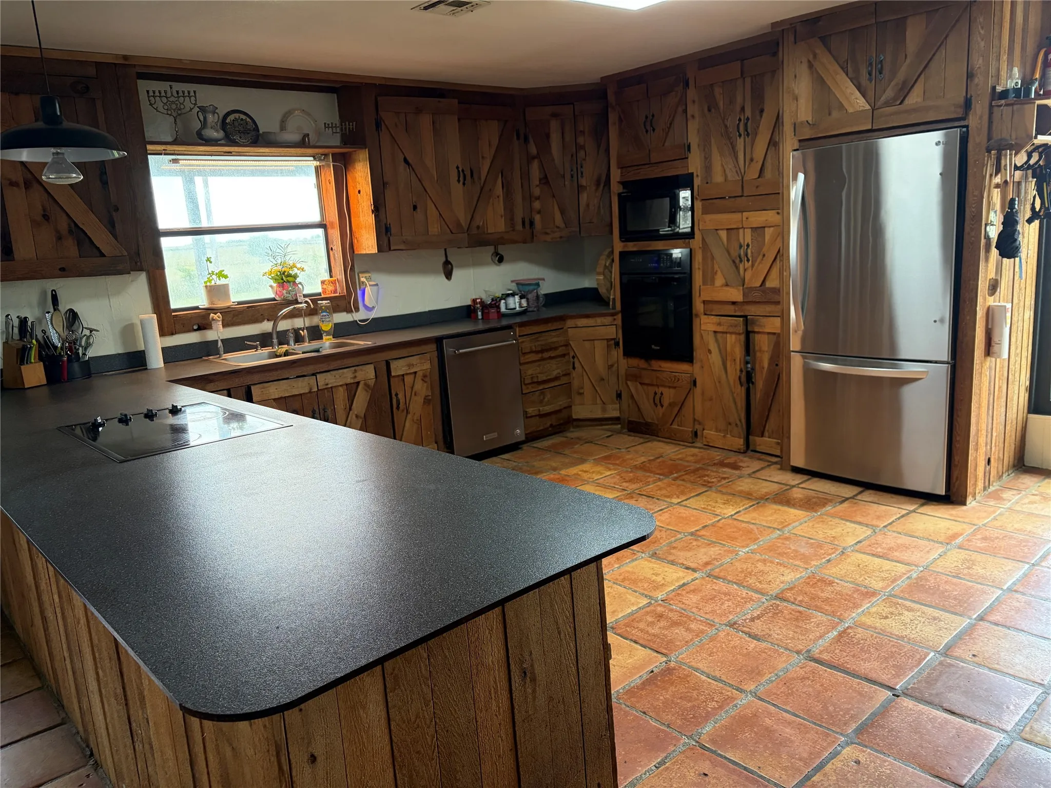 Kitchen featuring a large island with an integrated cooktop, stainless steel appliances, built-in microwave and oven, wood cabinetry with barn door detailing, and terracotta-style floor tiling