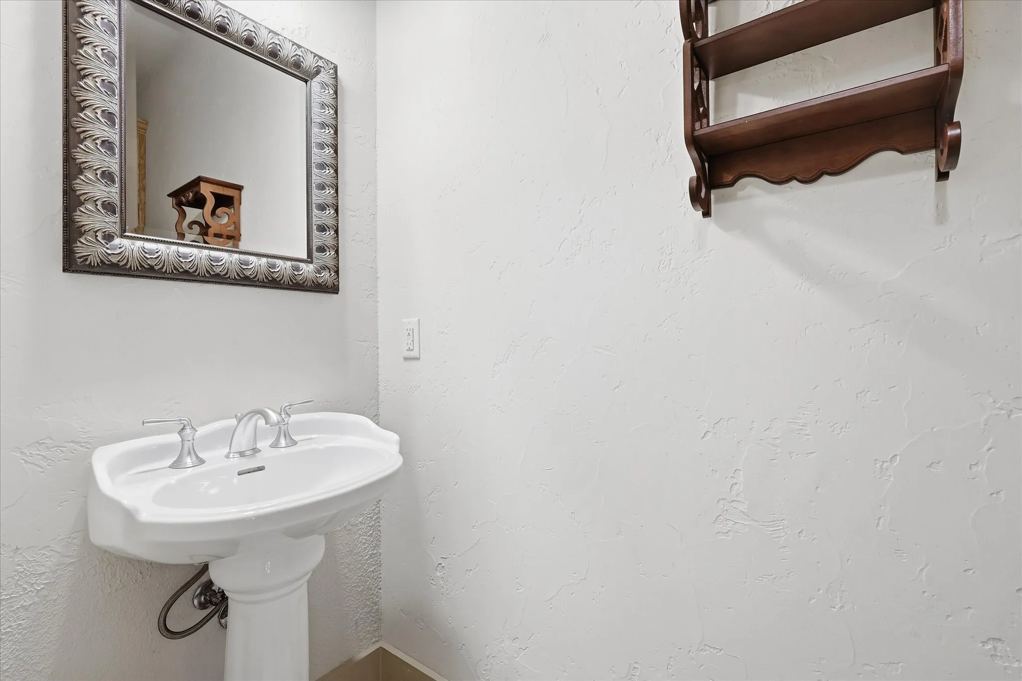 Pedestal sink with dual-handle chrome faucet, complemented by a framed mirror with an ornate surround