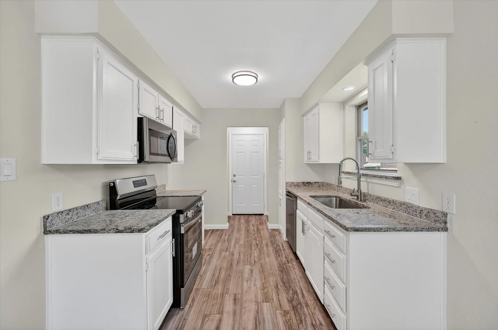 Galley kitchen featuring white cabinetry, granite-style countertops, wood-finish flooring, stainless steel appliances, and a window above the sink