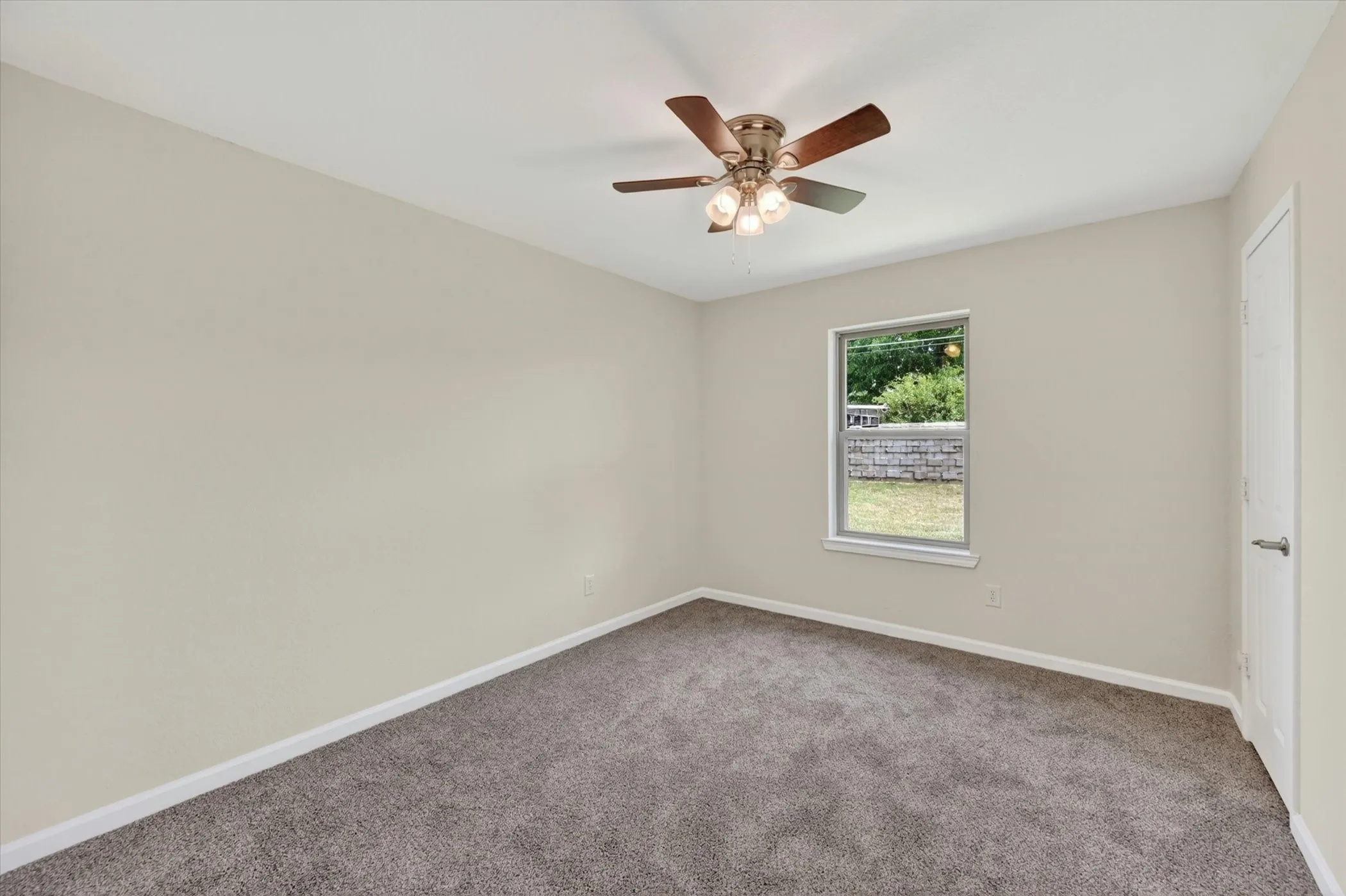 Spacious room featuring neutral wall paint, gray carpet, a single window, a ceiling fan with light fixture, and a white door with a brushed nickel handle
