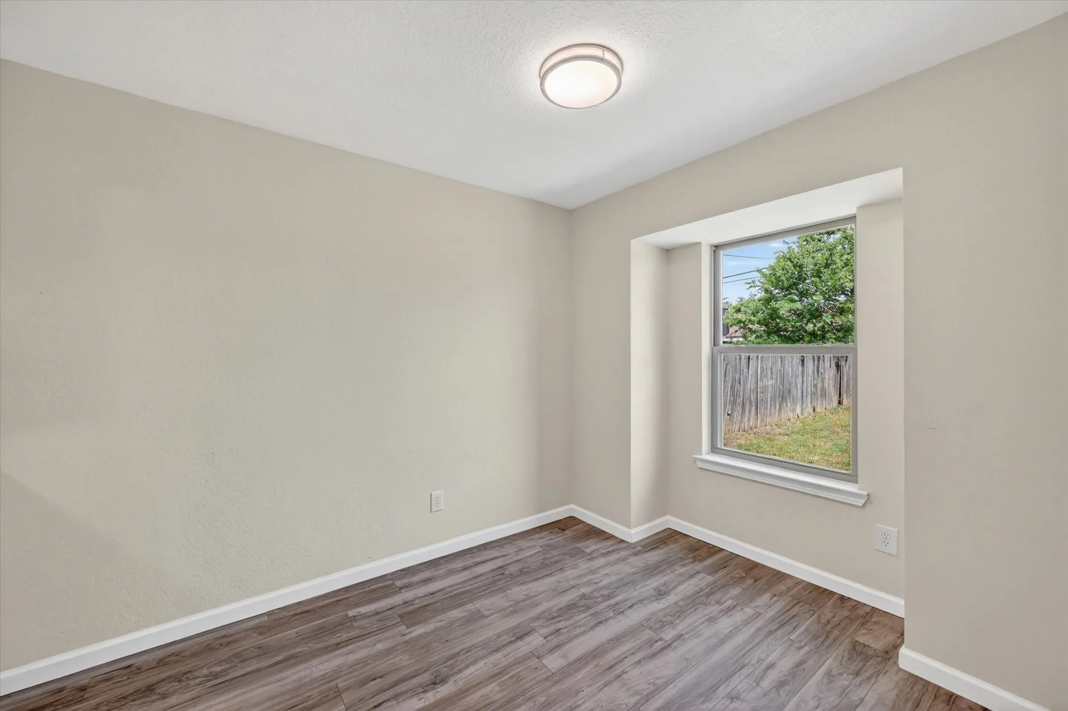 Interior room featuring wood-finish flooring, light-toned wall paint, a window with a white frame, and a flush-mount ceiling light fixture