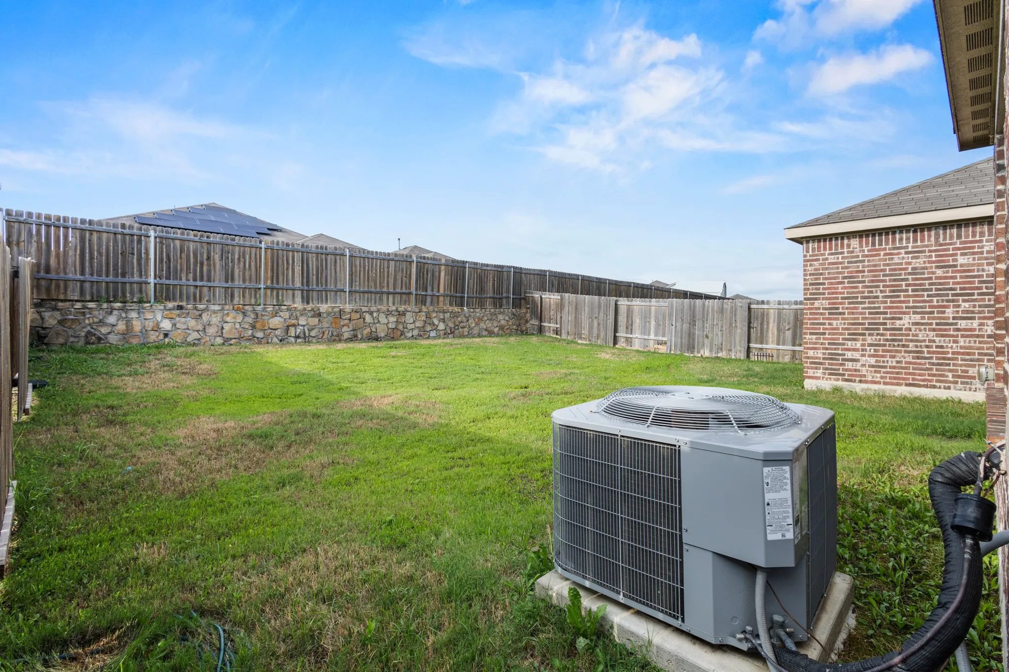 Spacious backyard featuring a full perimeter privacy fence with a stone base and wood paneling, a brick exterior wall, and an air conditioning unit on a concrete pad