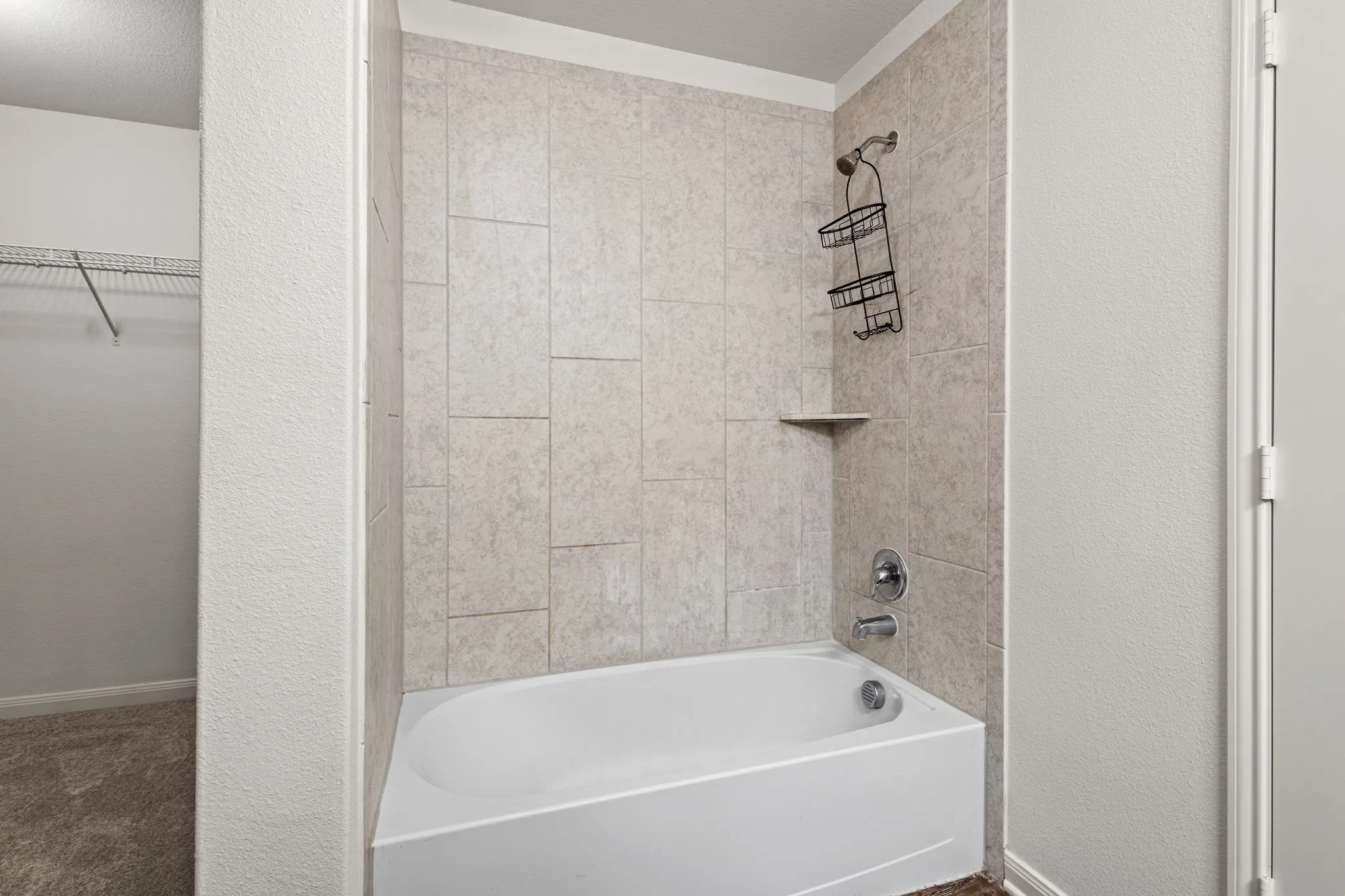 Full tub and shower combination featuring neutral-toned wall tiling, chrome fixtures, and a built-in corner shelf