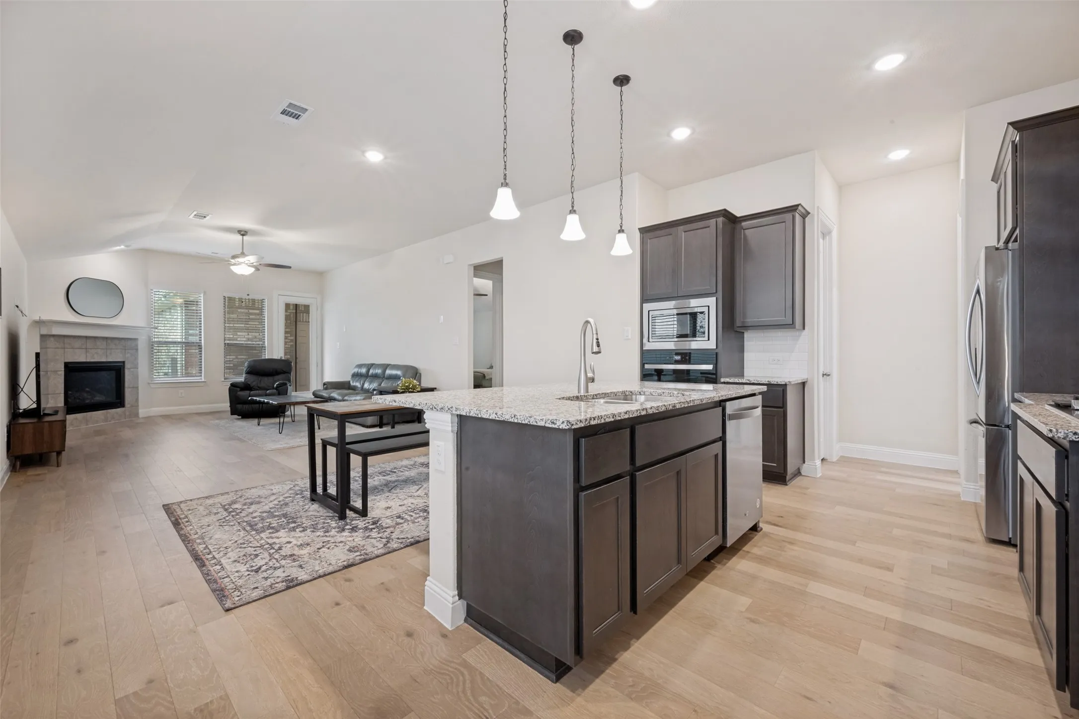 Open-concept kitchen featuring an island with sink and pendant lighting, granite countertops, stainless steel appliances, and dark wood cabinetry
