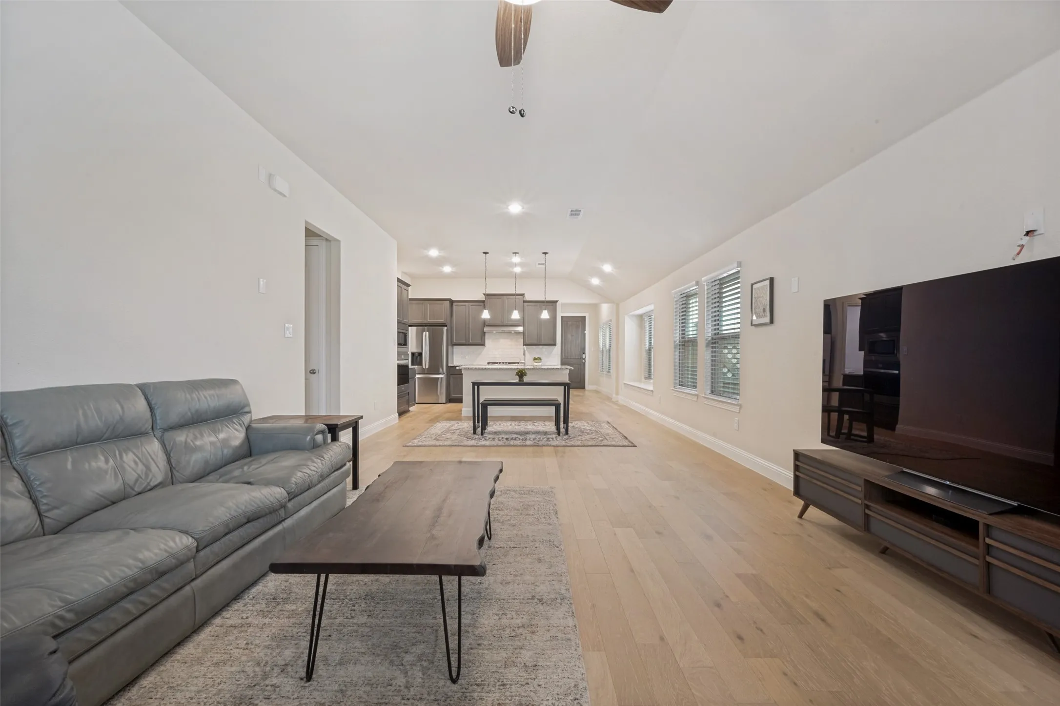 Open-concept living area featuring light wood-finish flooring, recessed lighting, vaulted ceiling, a row of windows with blinds, and a kitchen visible in the background