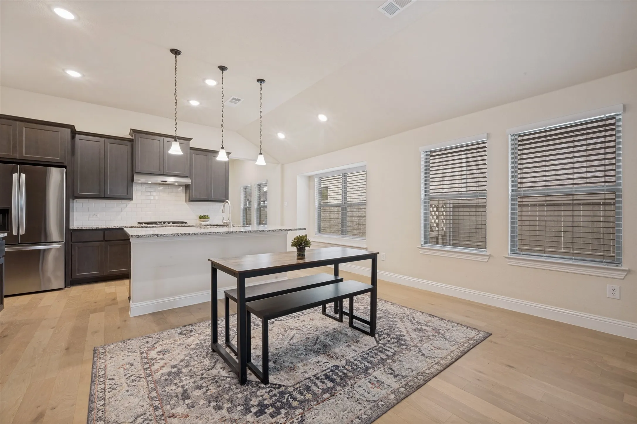 Open-concept kitchen and dining area featuring light wood-finish flooring, dark cabinetry, stainless steel appliances, and a white subway tile backsplash