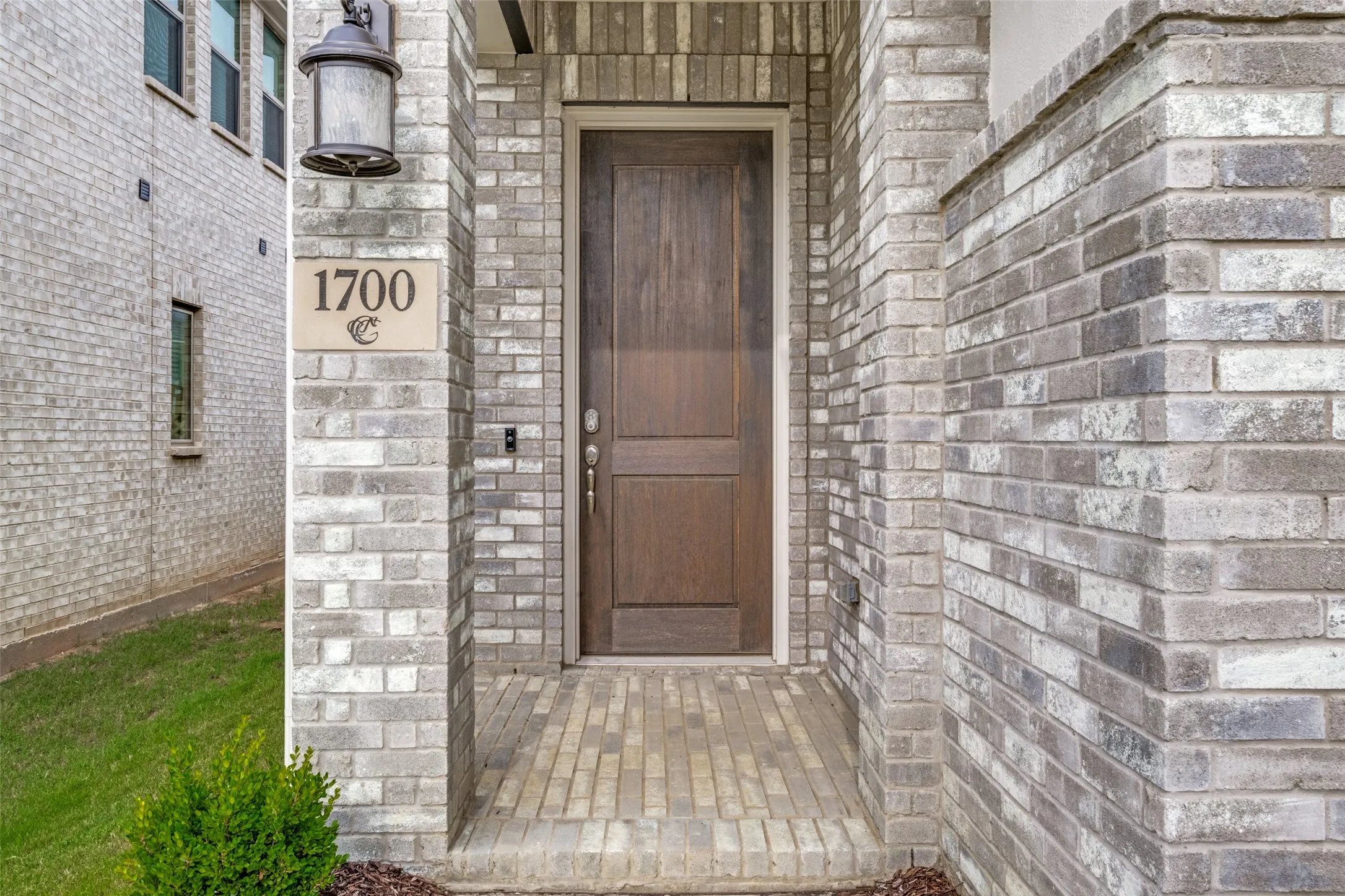 Entryway featuring a wood-panel door, varied gray brick facade, exterior sconce lighting, and a paver landing