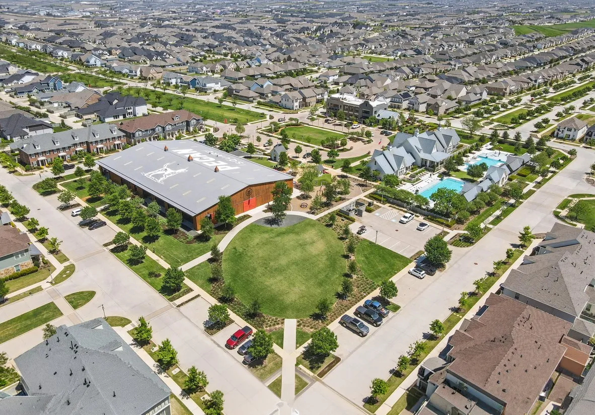 Expansive community featuring multiple residential streets with single-family homes and townhomes, a large recreational building with a metal roof, a prominent community pool area with multiple pools, and a central green space with walking paths