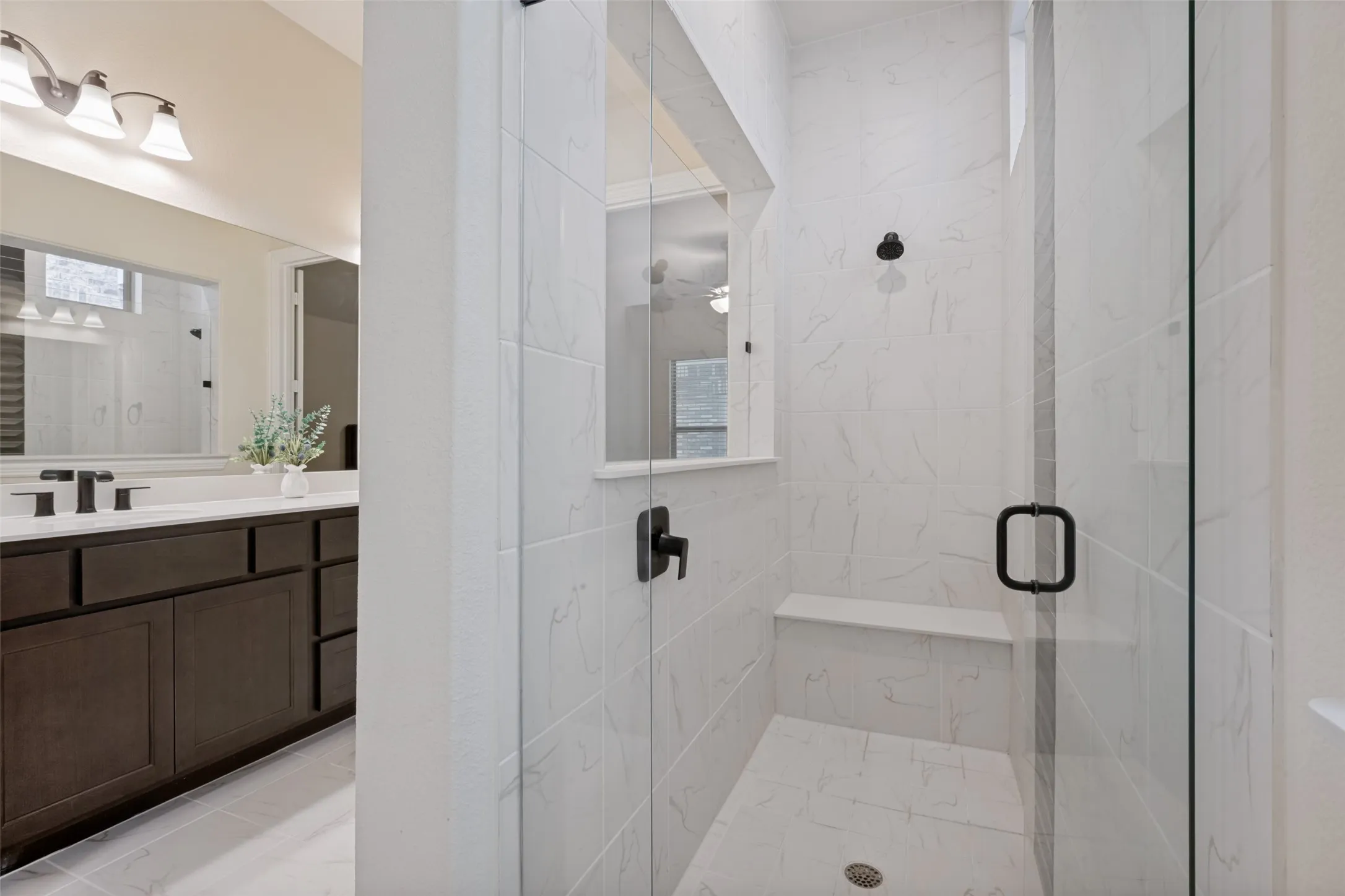 Walk-in shower with clear glass enclosure and black hardware, featuring white tiled walls and flooring, a built-in shower bench, and a matte black rainfall showerhead