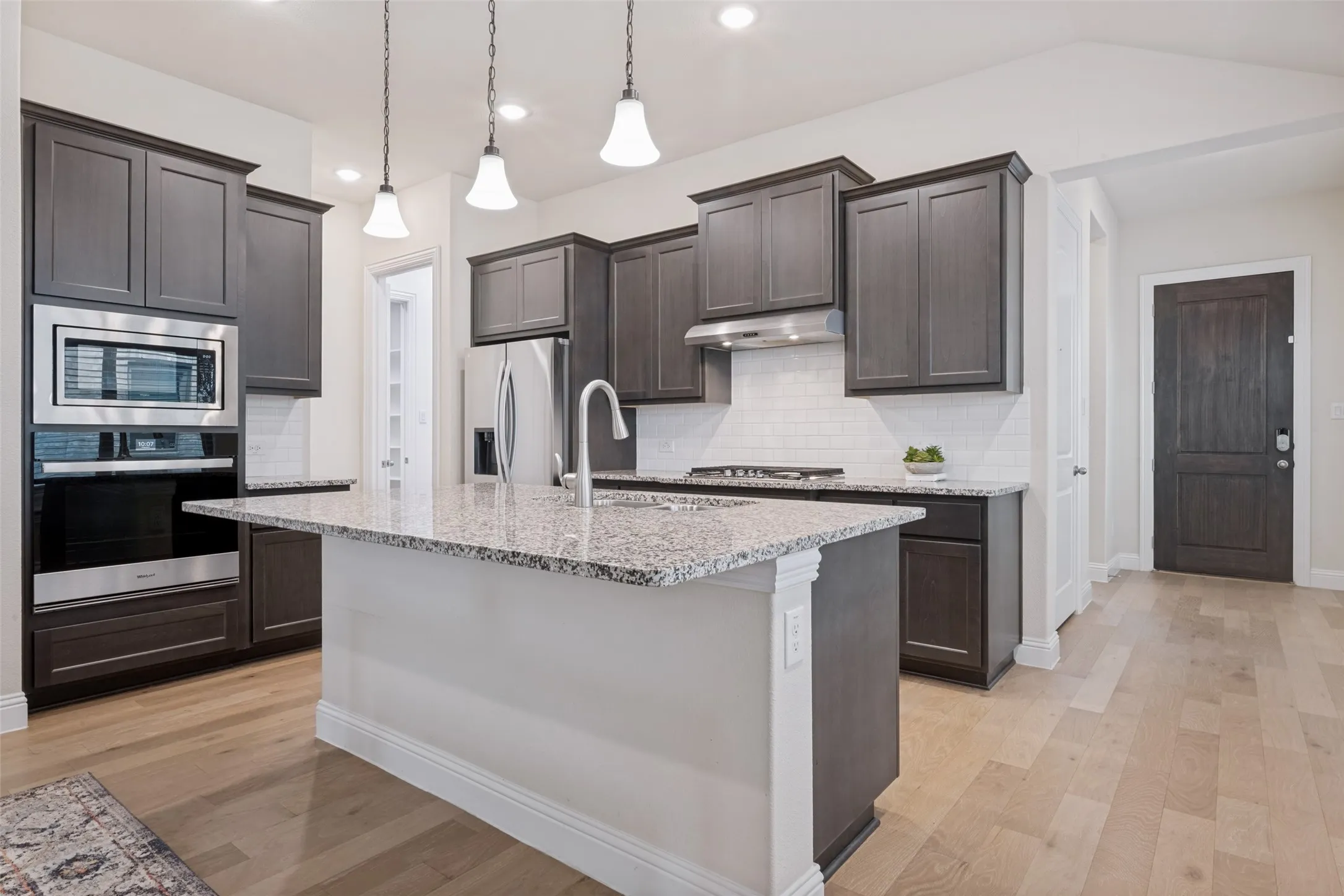 Kitchen featuring a central island with granite countertop, stainless steel appliances, dark wood-finish cabinetry, white subway tile backsplash, and light wood-finish flooring