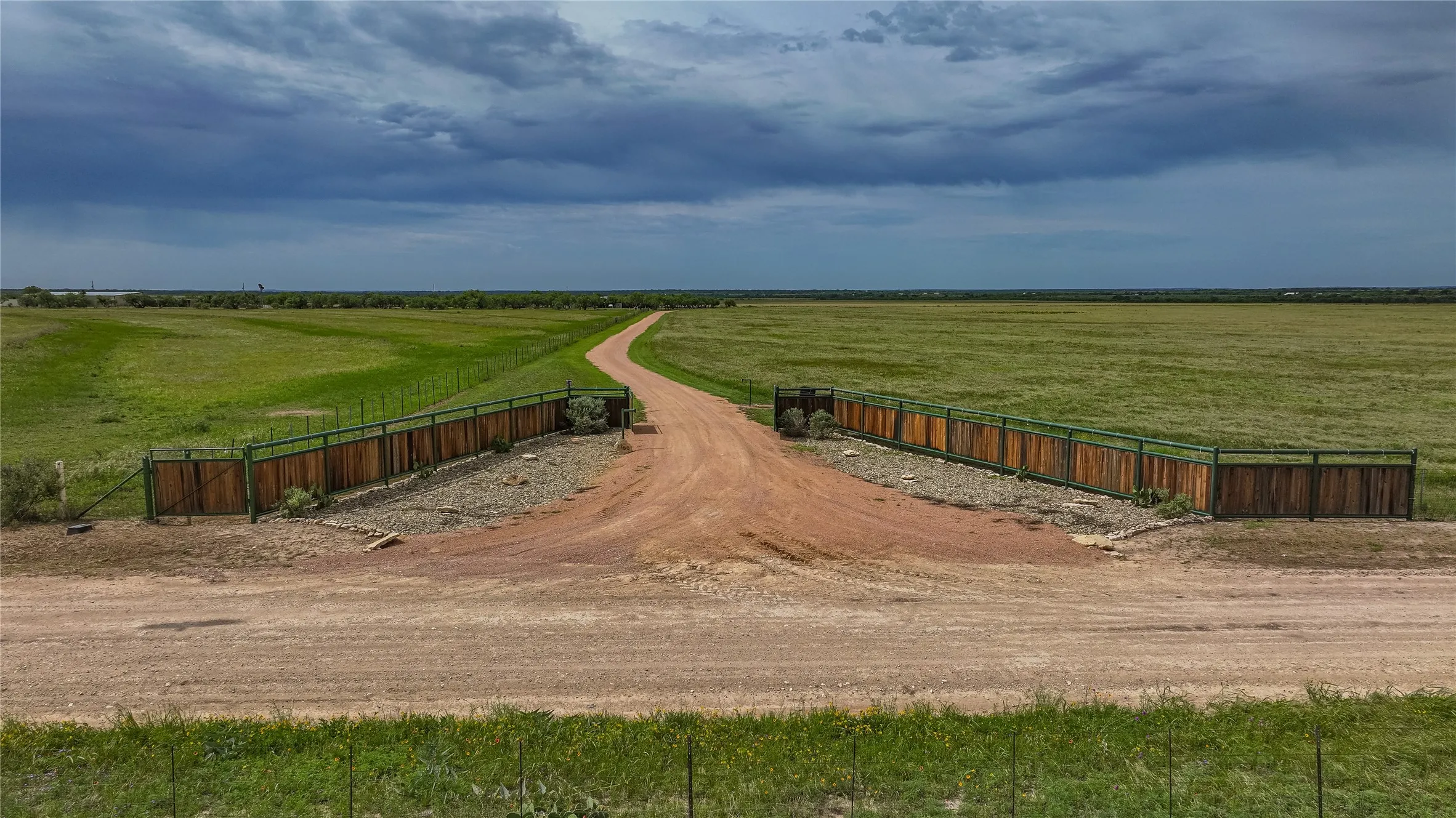 Private ranch entrance on CR 419 with custom steel and cedar entry fencing. A wide caliche drive leads through open coastal Bermuda pasture, ideal for a cattle ranch, hay operation or horse property.