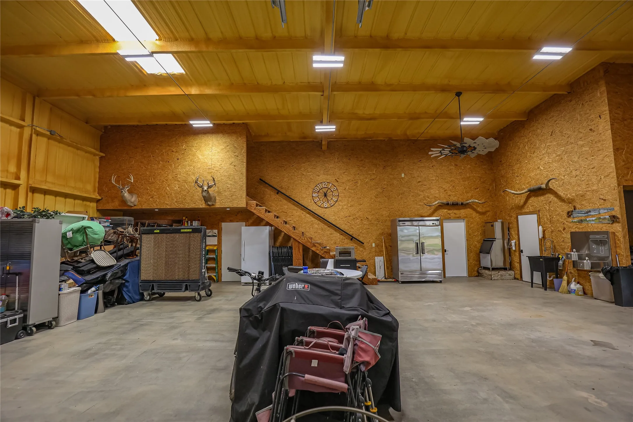 Insulated shop and gathering space with a soaring ceiling, roll up door, commercial ice machine and stainless refrigerator. Whitetail mounts on the wall make it a natural hunting lodge or game room.
