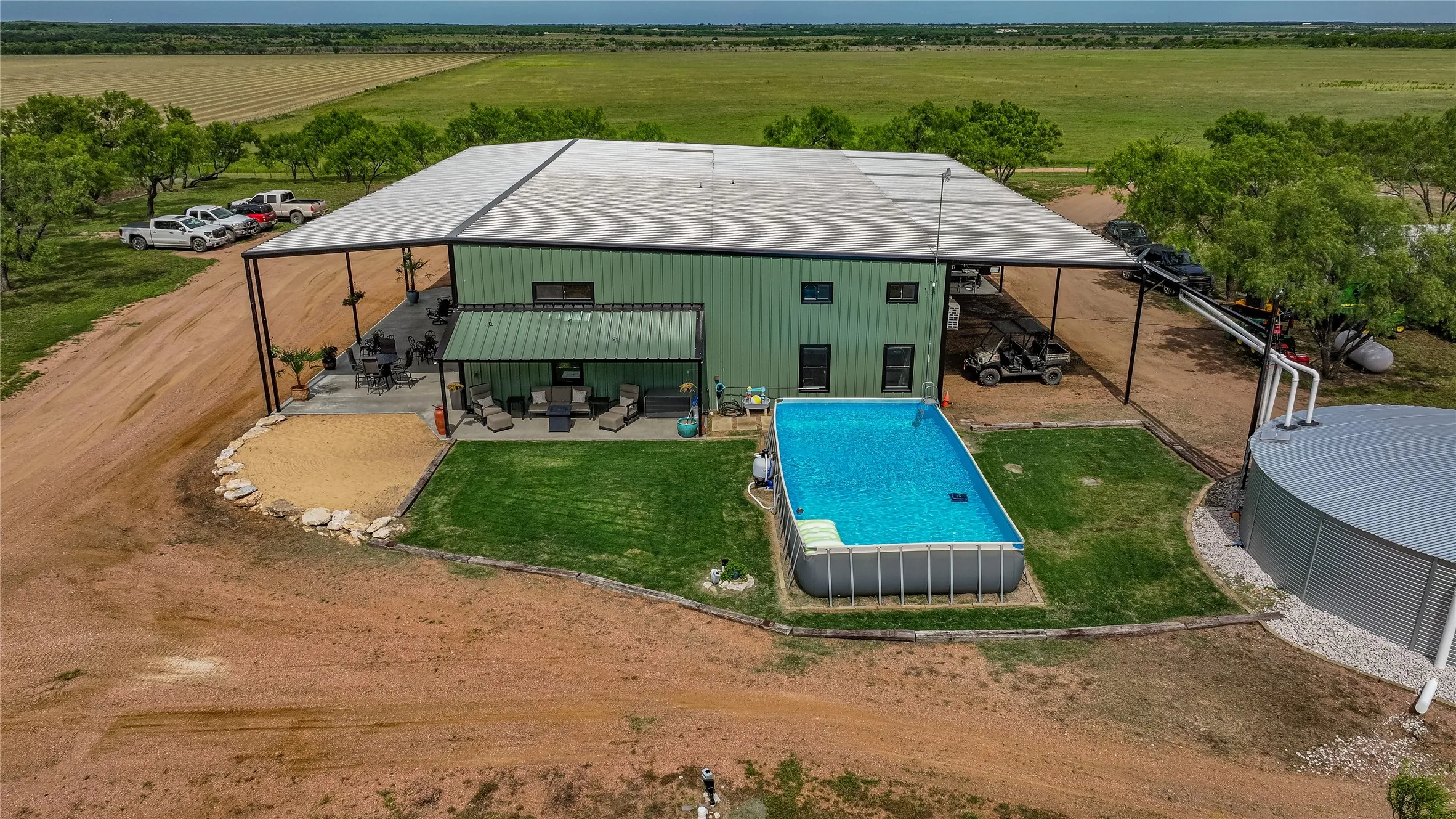 Aerial of the barndominium backyard: a fenced lawn, above ground pool, covered patio and attached carport parking. Productive hay fields and native mesquite stretch beyond.