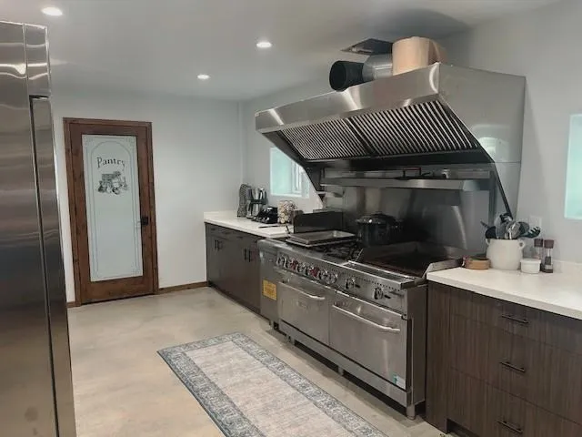 Commercial kitchen with a six burner range, griddle, double ovens, fryer and a stainless vent hood. A two door reach in refrigerator and pantry shelving round it out, ready for hunting lodge meals, retreat catering or event venue use.
